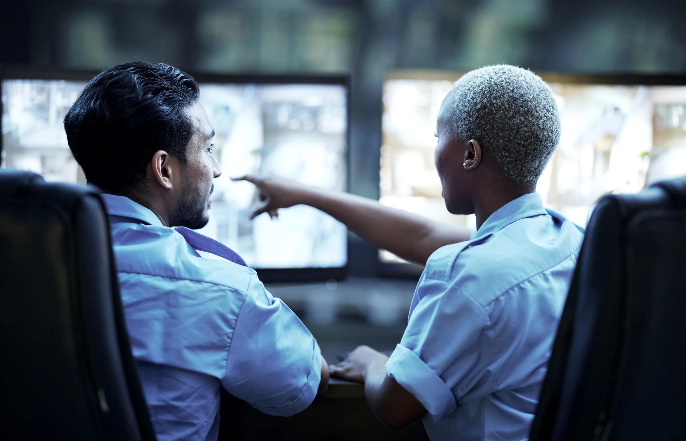 Two security operators in light blue shirts seated at a desk, one pointing at a wall of camera monitors