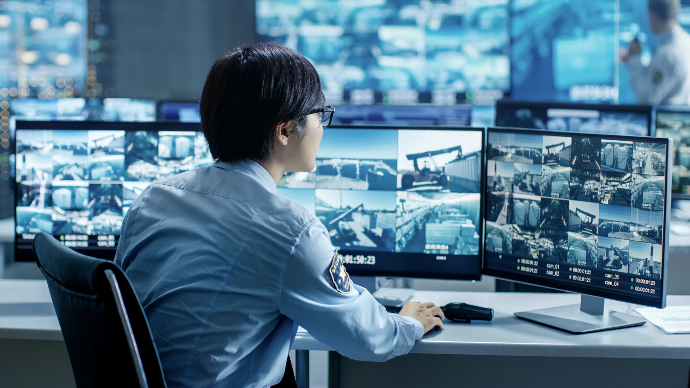 Operator in a light blue shirt seated at a control desk facing a wall of camera monitors