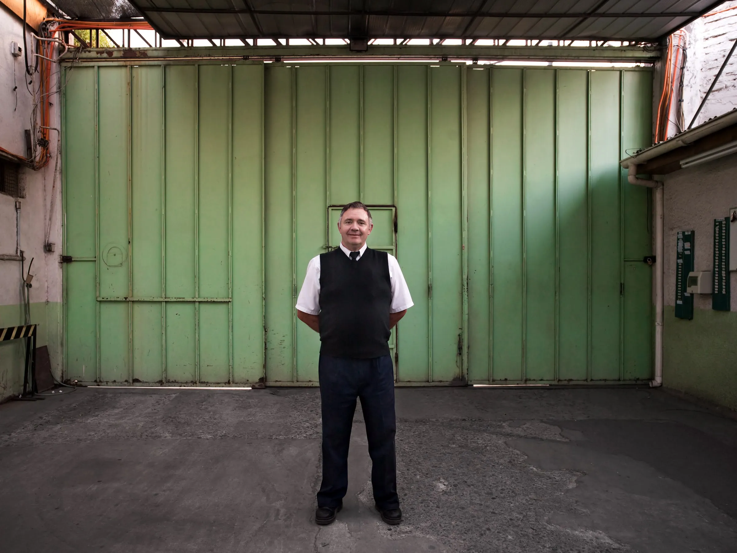 Security guard in a dark vest standing with hands clasped in front of a large green warehouse door