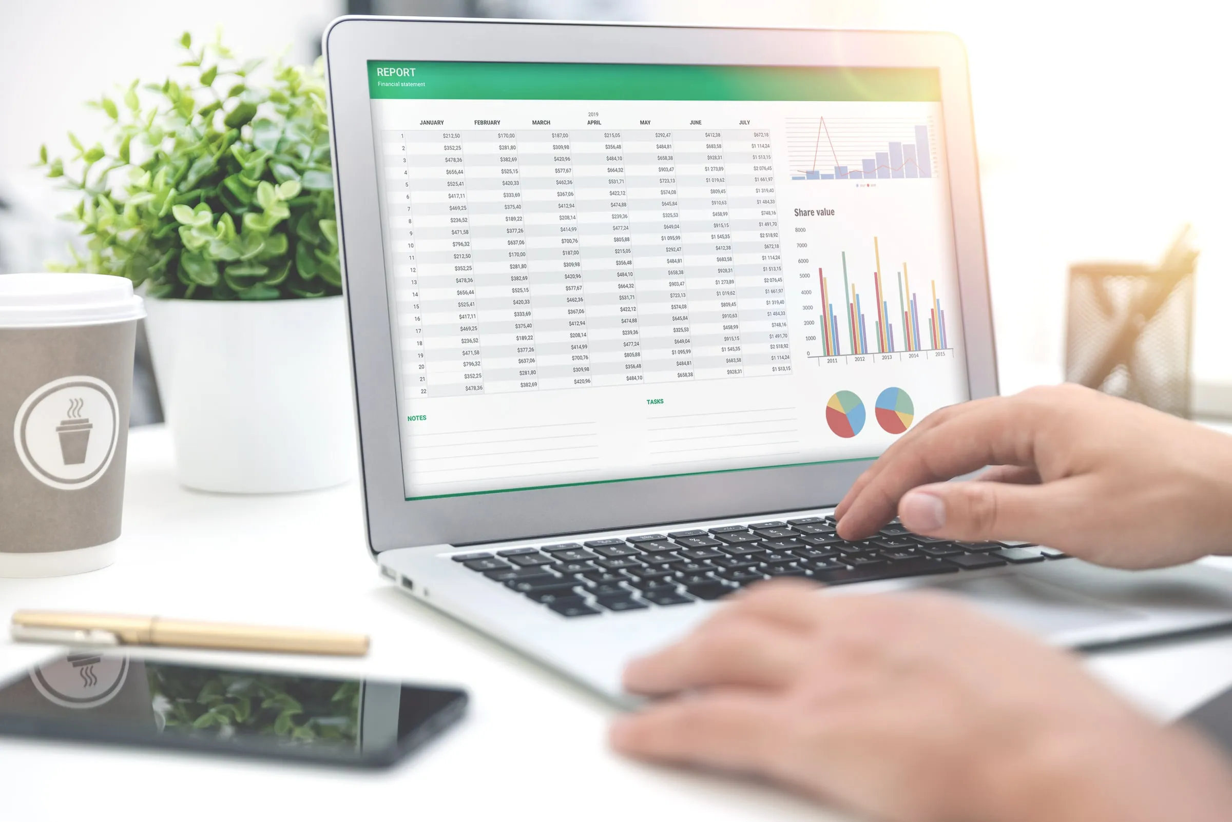 Hands typing on a laptop showing a spreadsheet, beside a coffee cup and a small potted plant on a white desk
