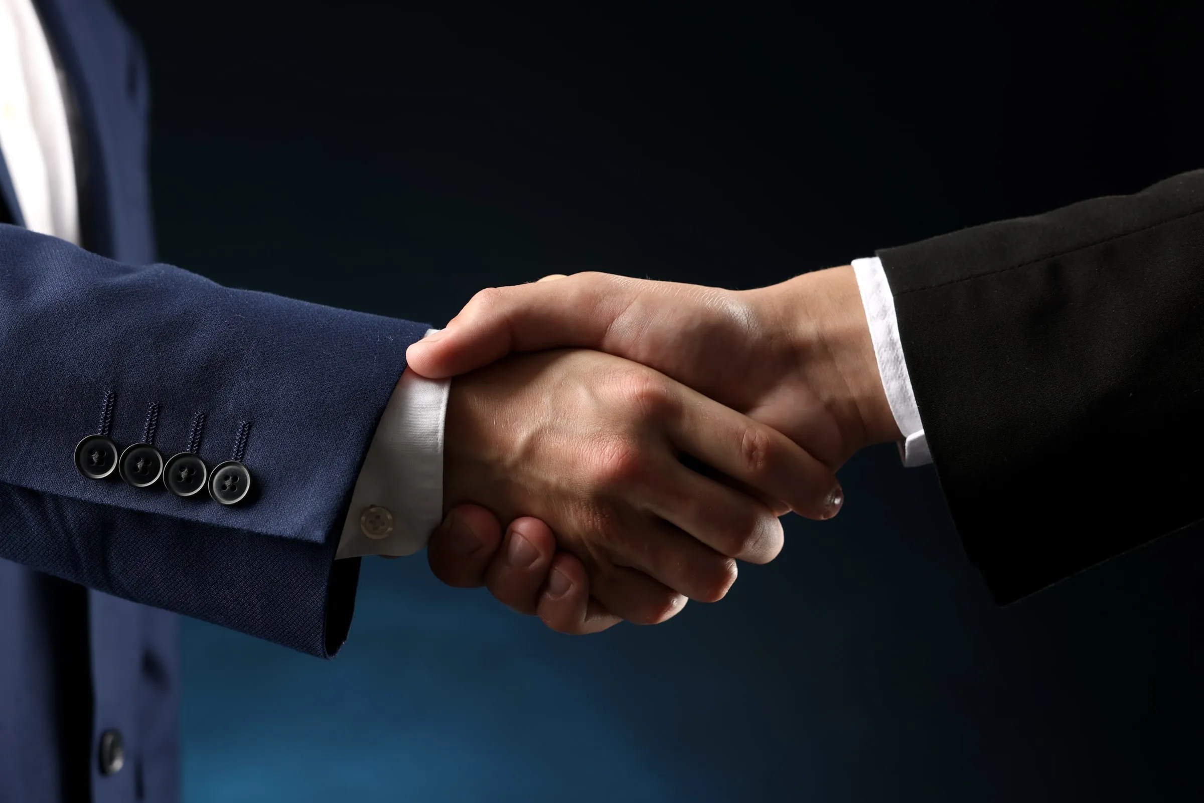 Two people in suits shaking hands, close-up on the handshake against a dark background
