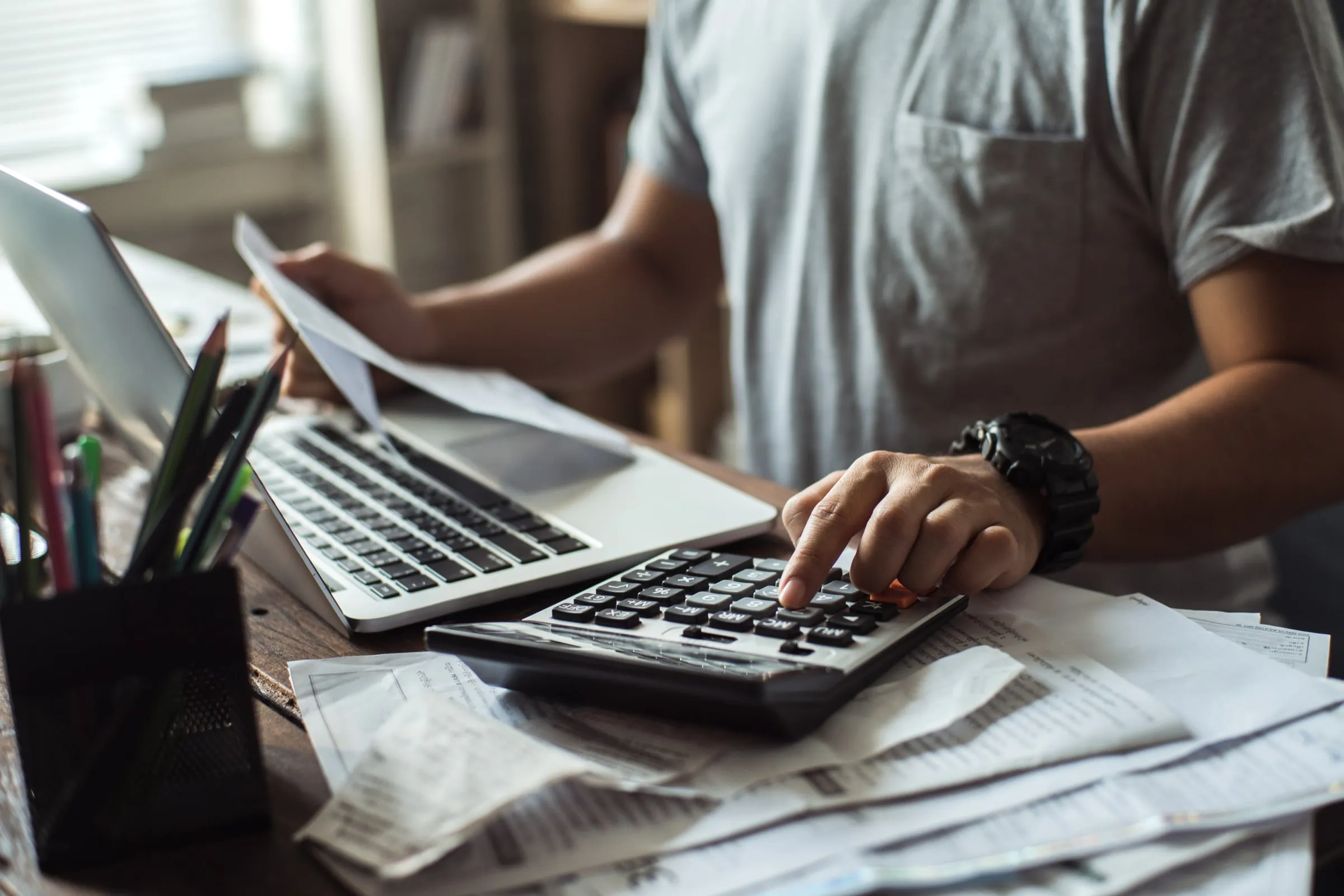 Person at a desk pressing calculator keys beside a laptop, papers and a coffee cup