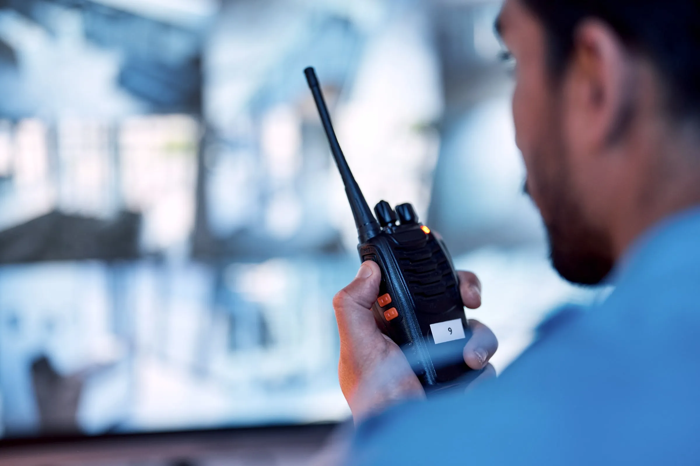 Security officer in a blue uniform holding a handheld radio, with a wall of camera feeds blurred behind