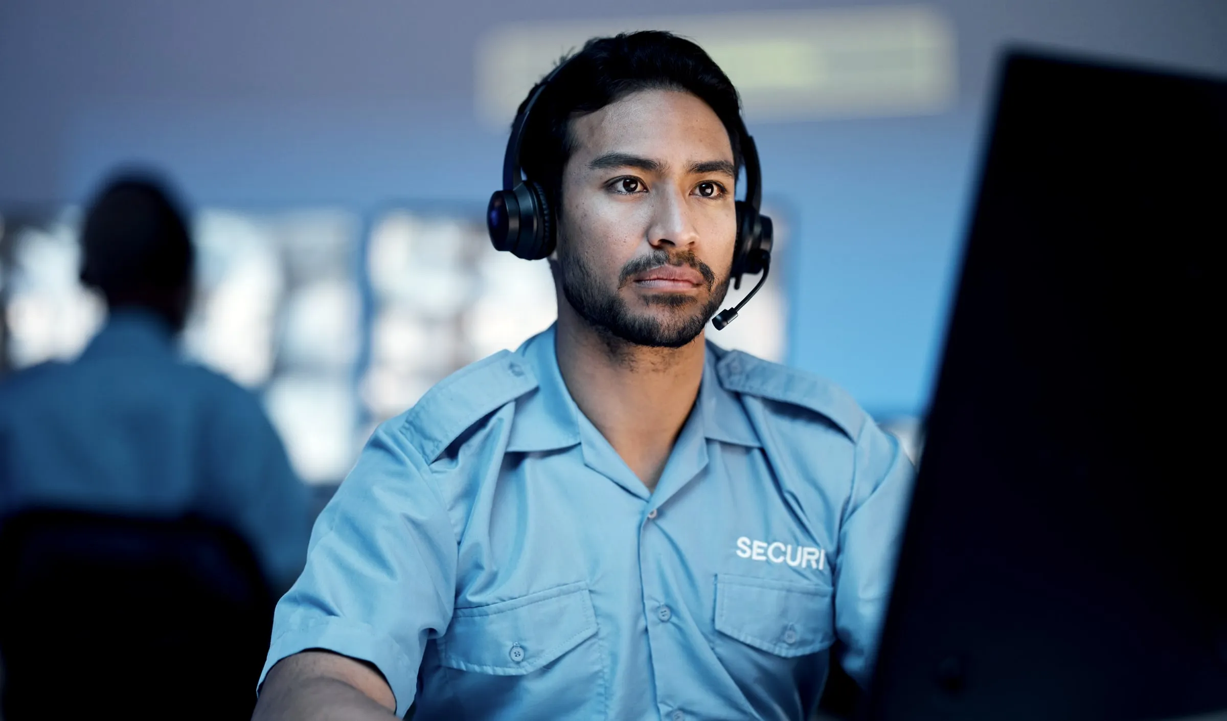 Security officer in a blue SECURITY shirt wearing a headset, seated at a monitor in a control room