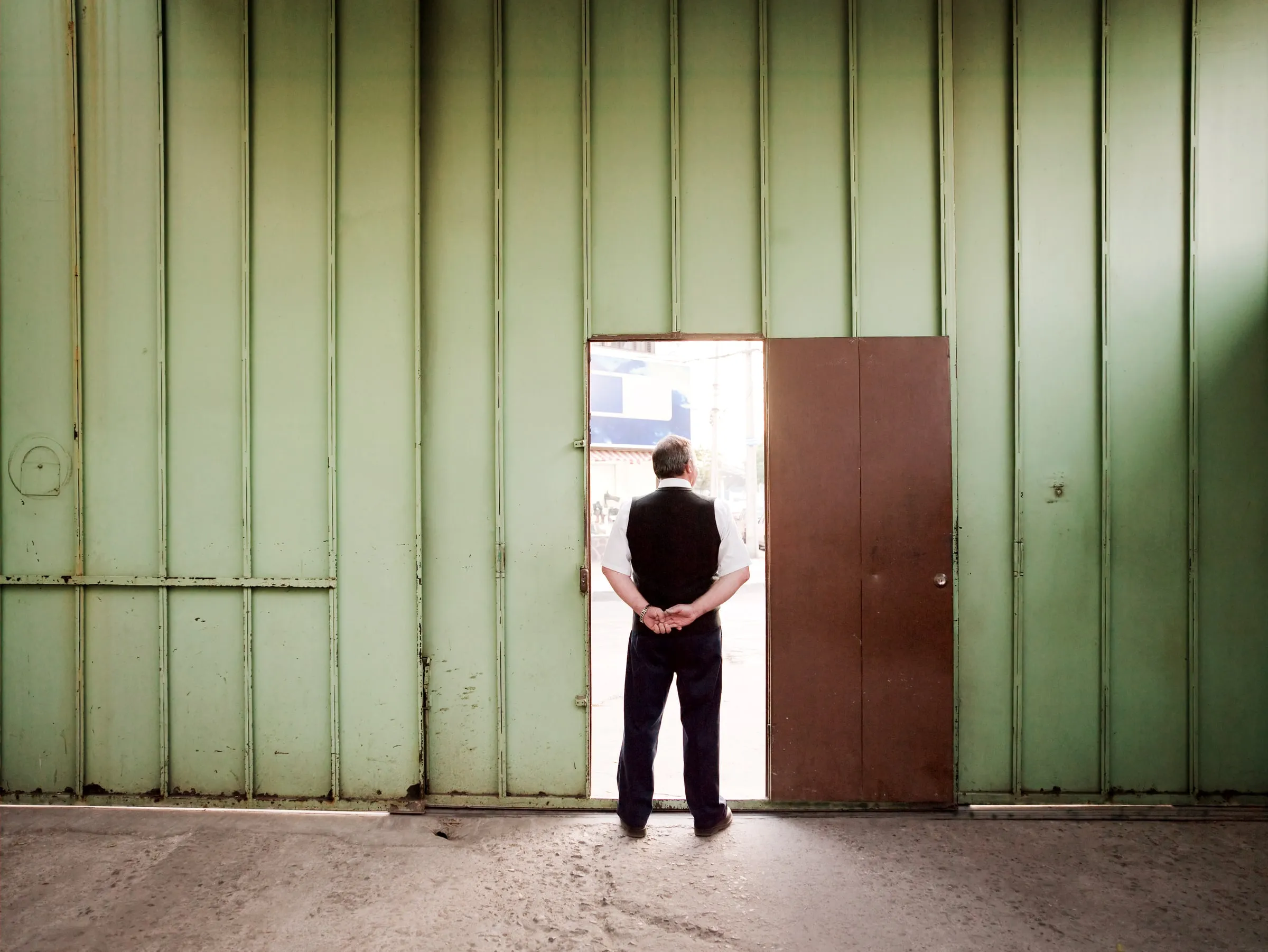 Security guard in a dark vest standing in an open doorway of a green warehouse wall, seen from behind