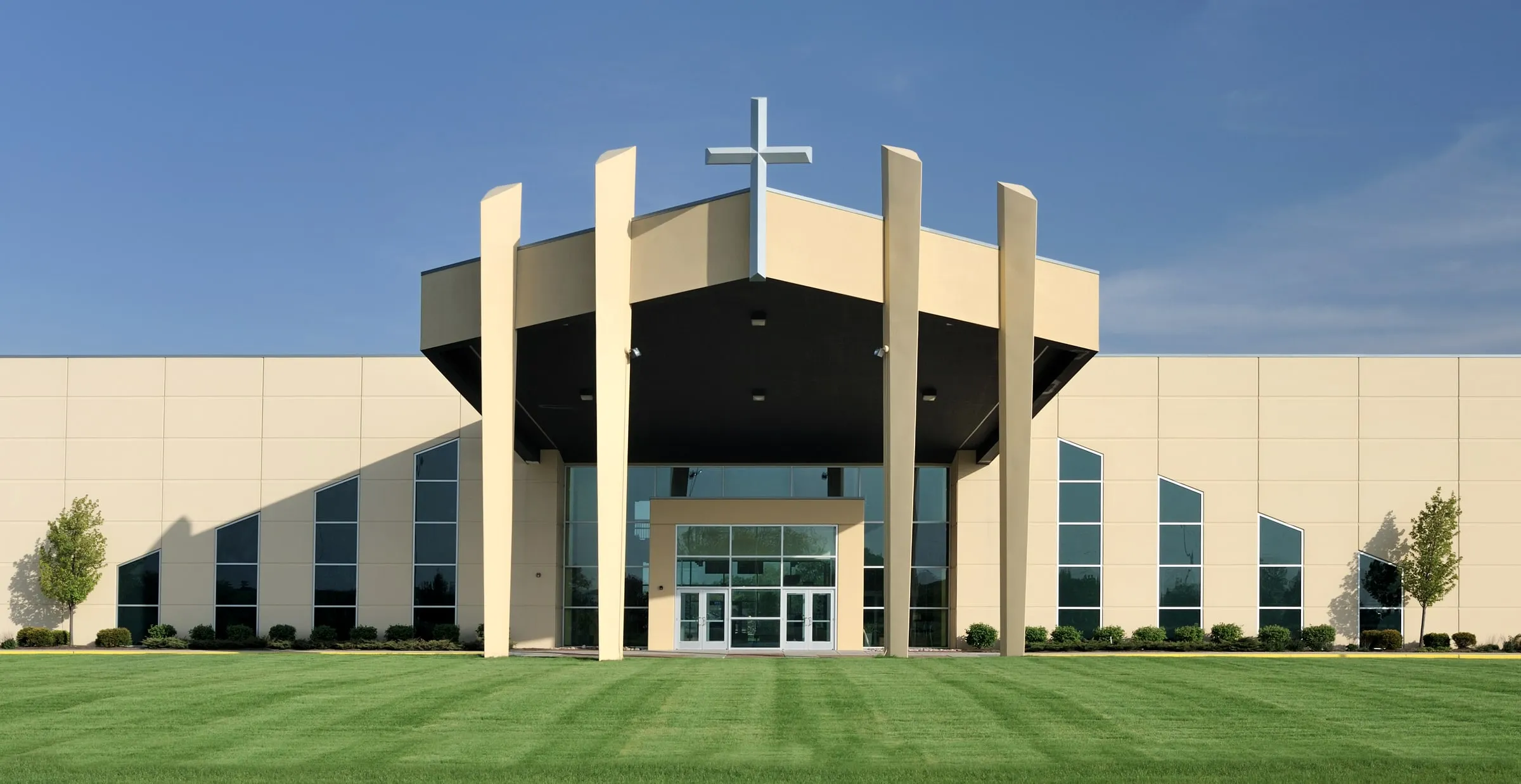 Modern church with a cross on the roof, tall windows and a wide green lawn under a blue sky