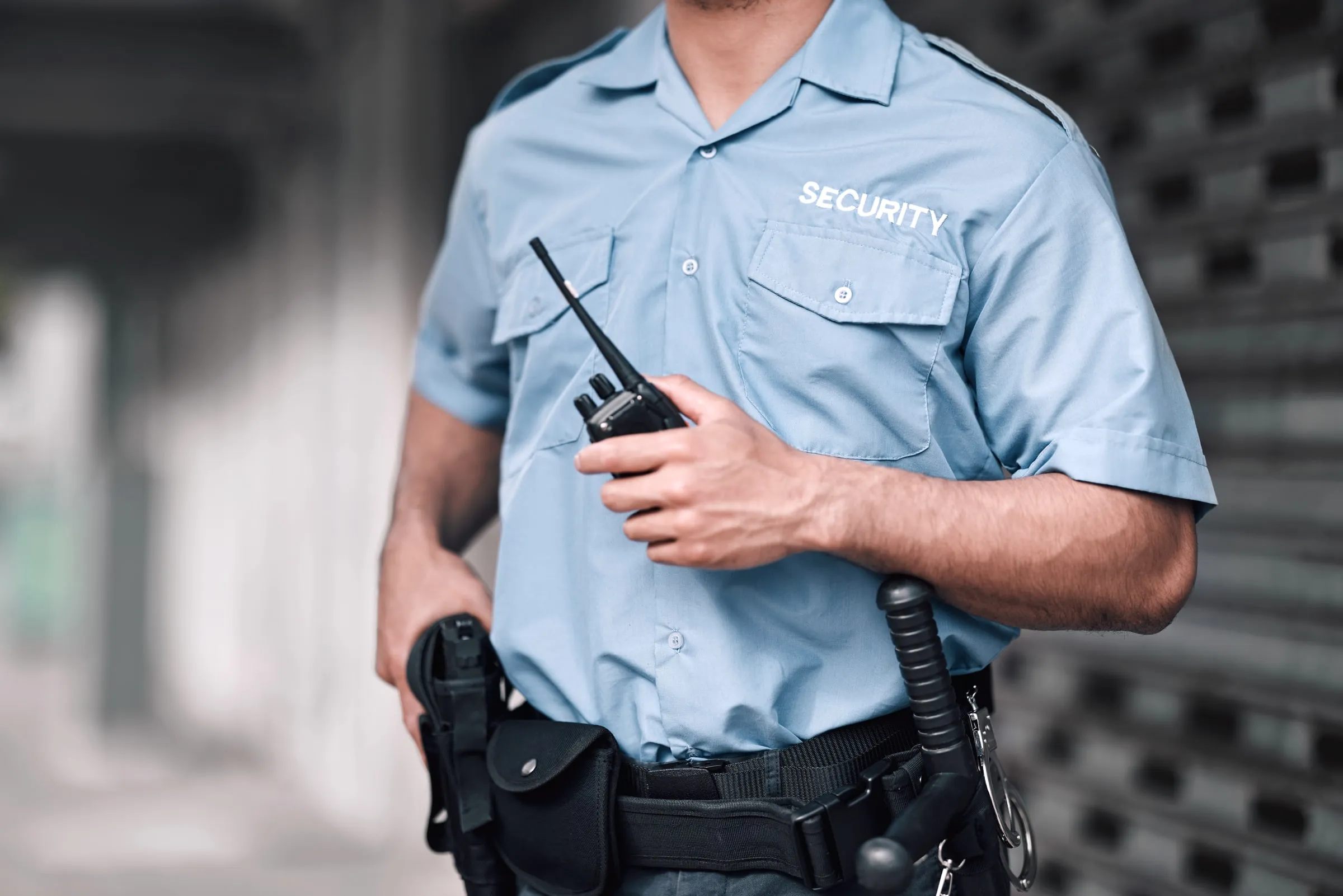 Security officer in a light blue shirt holding a radio, with a duty belt and handcuffs, seen from the chest down