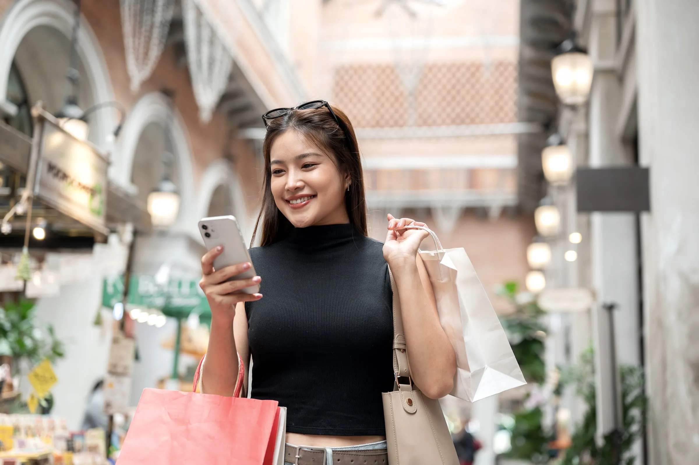 Smiling shopper with sunglasses on her head carrying shopping bags and looking at her phone on a retail street