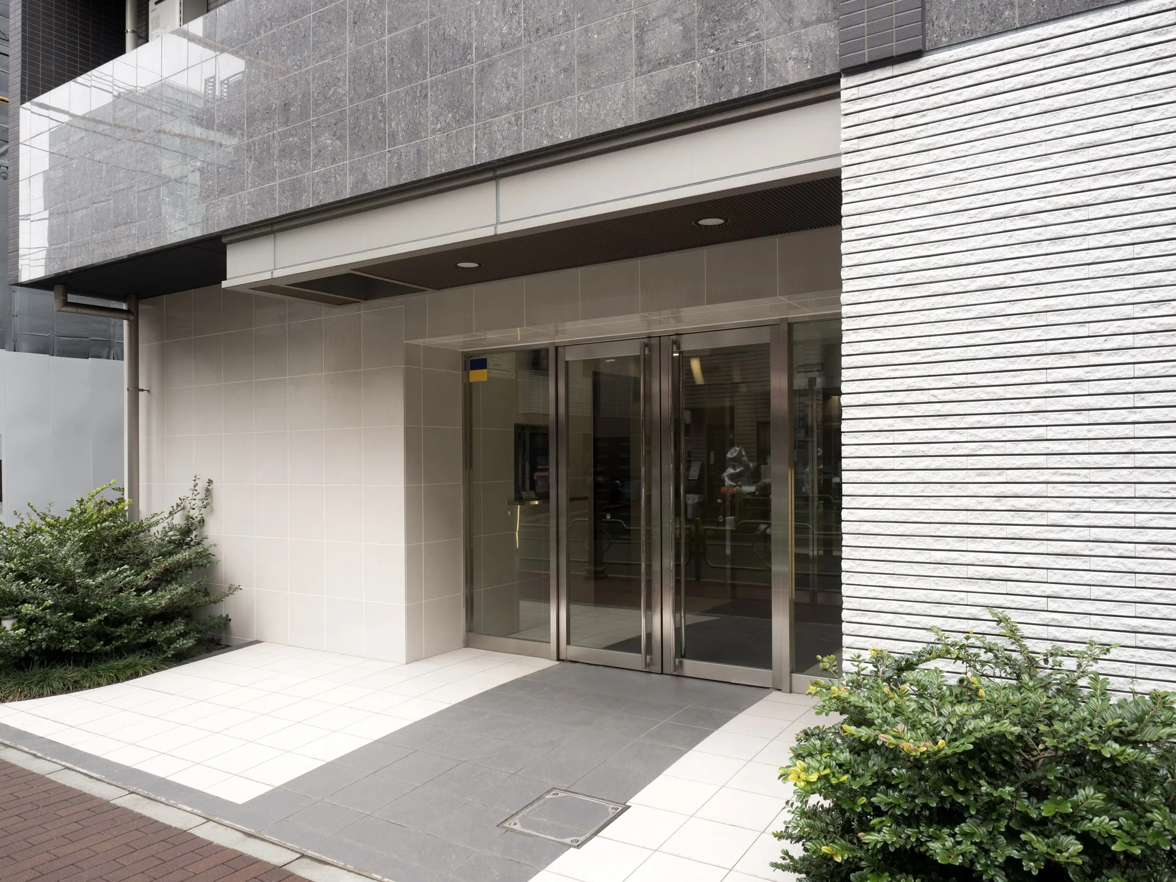 Glass entrance doors of a modern building with a tiled walkway and planted border