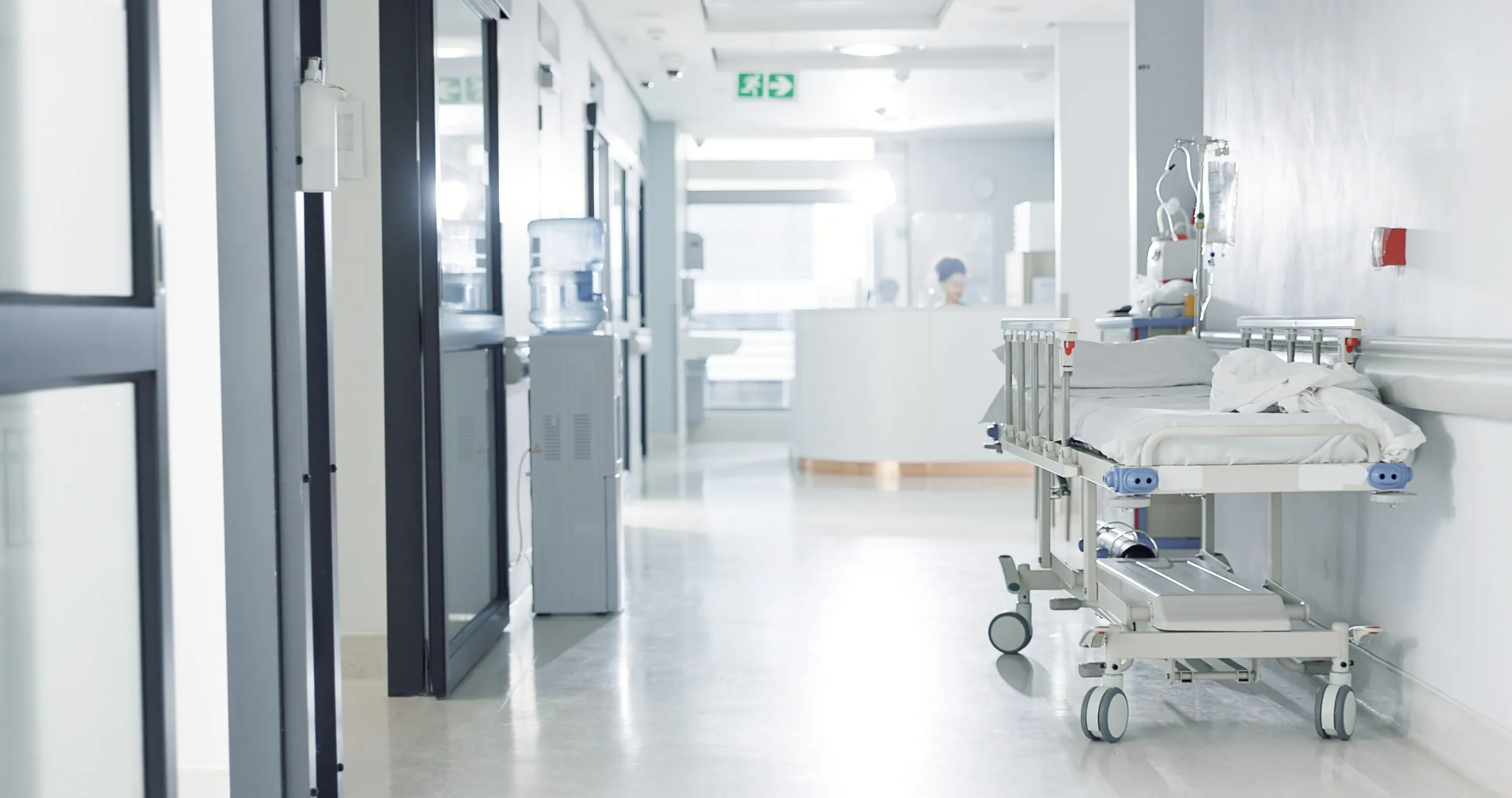 Bright hospital corridor with an empty gurney, wall rails and staff in scrubs at the far end