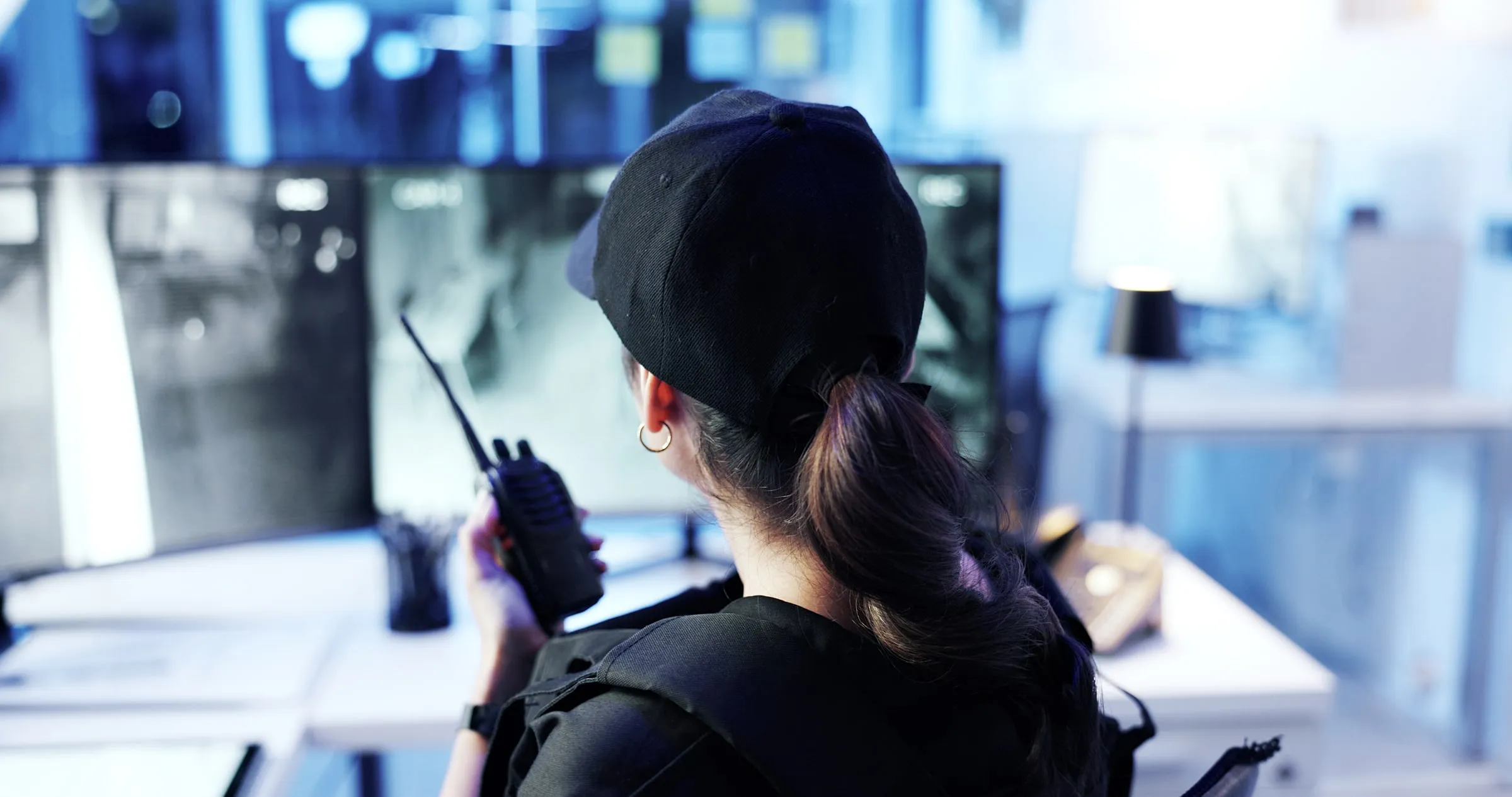 Female security officer with a radio and a dark vest at a desk in front of camera monitors, seen from behind