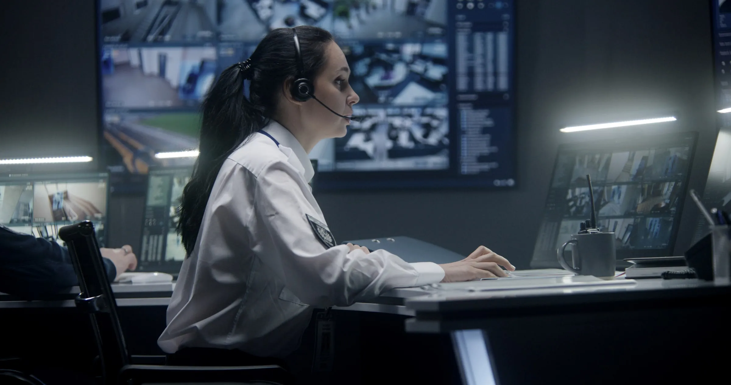 Operator wearing a headset at a desk facing camera monitors in a dimly lit control room