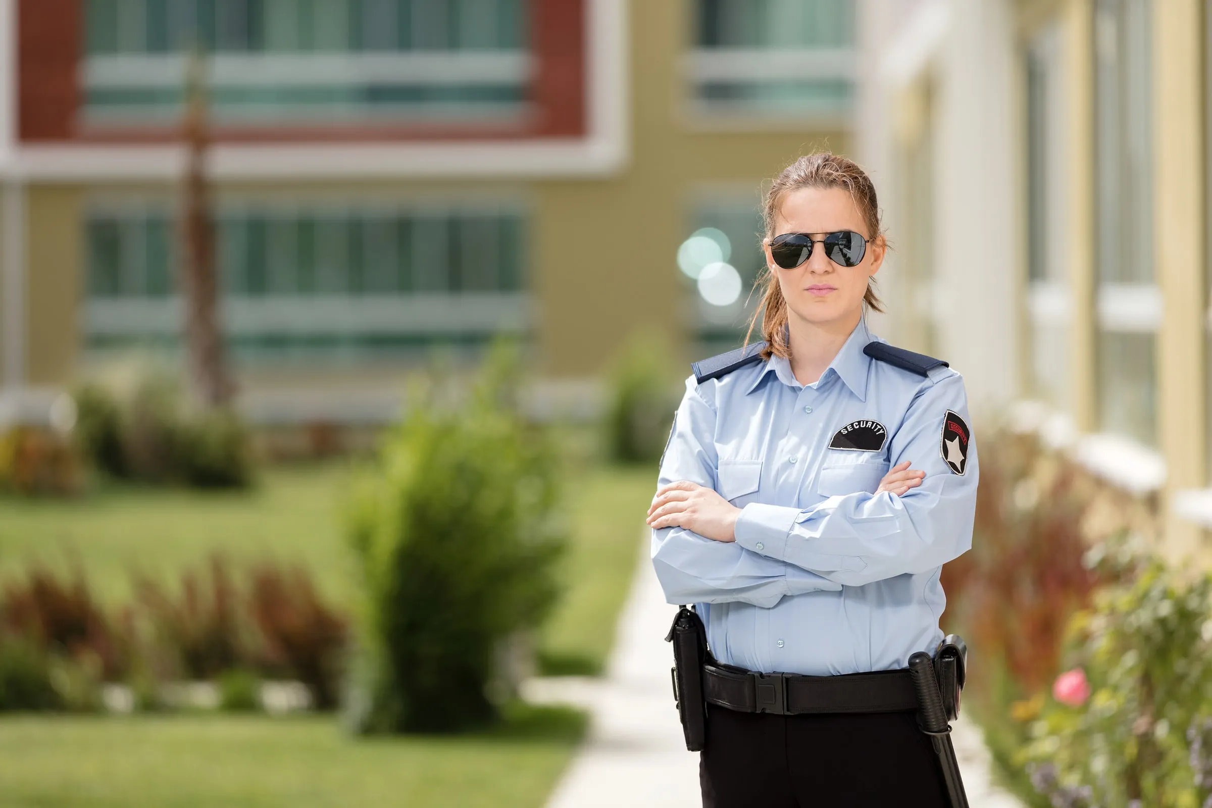 Female security officer in a light blue uniform and sunglasses standing with arms crossed outside a building