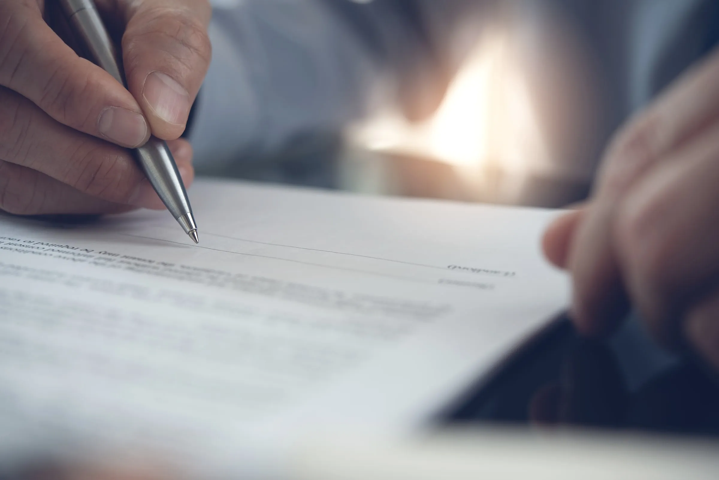 Close-up of a hand holding a pen over a printed document, with a second person's hand at the edge of the page
