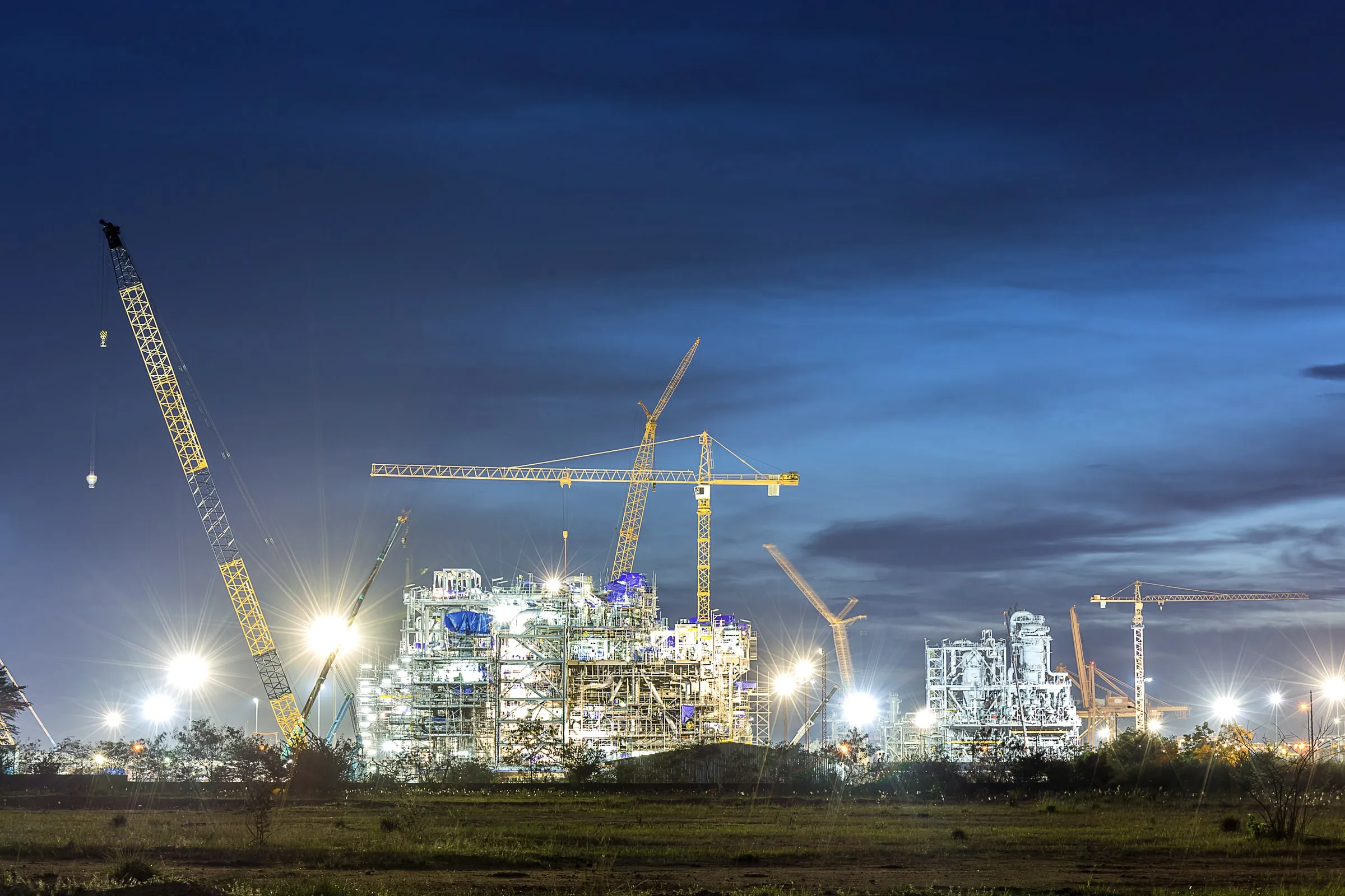 Construction site at dusk with tower cranes and floodlights against a darkening sky