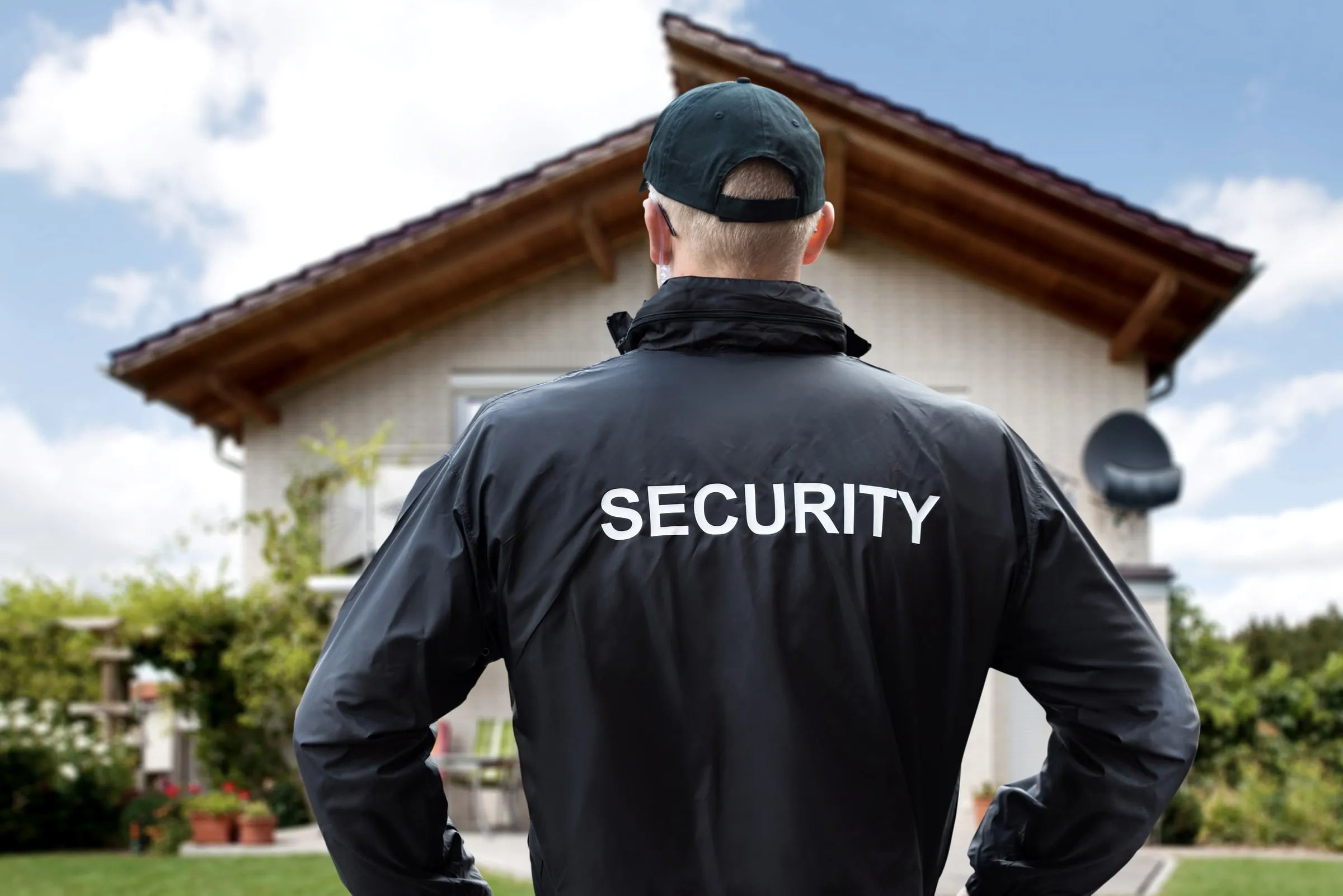 Security officer with SECURITY on the back of a black jacket, standing on a lawn in front of a house with a porch