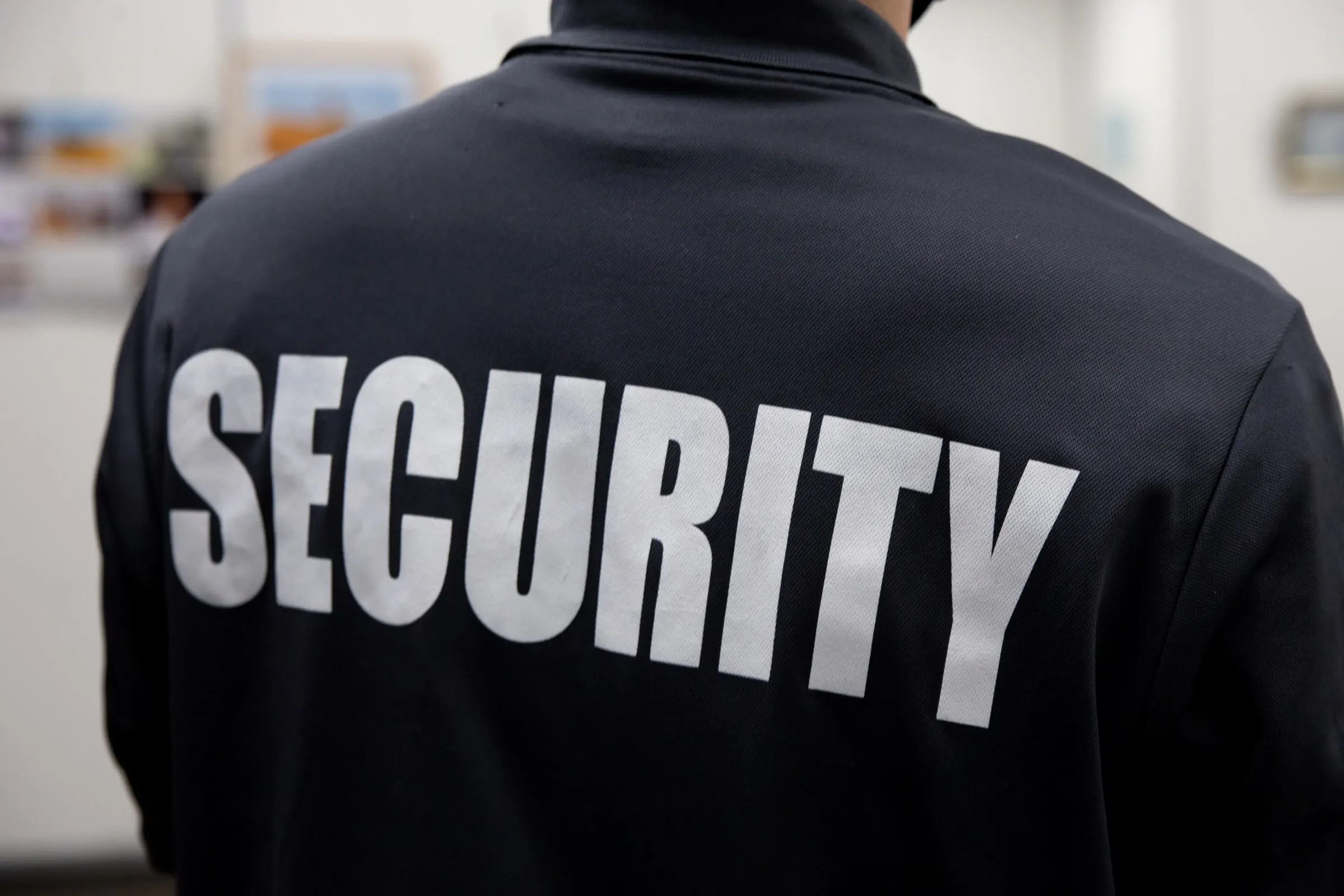 Back of a security officer in a black jacket with SECURITY in white letters, in a dim hallway