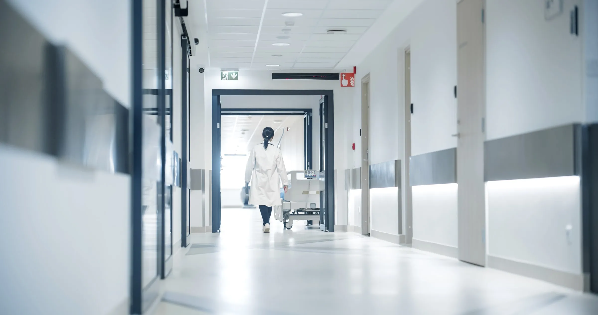 Long bright hospital corridor with a nurse walking away and a wheelchair against the wall
