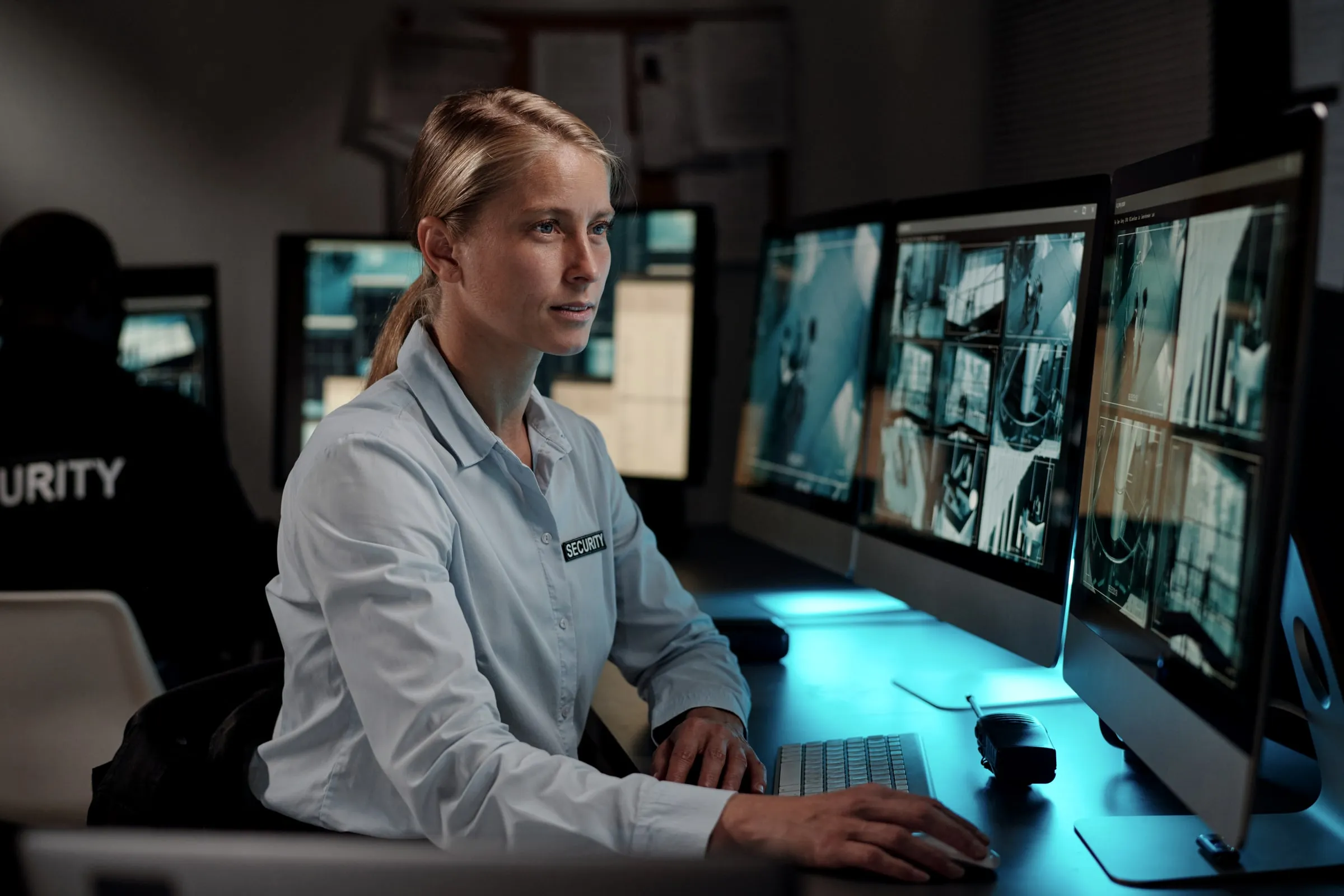 Female security operator in a light shirt at a desk with two monitors showing camera feeds