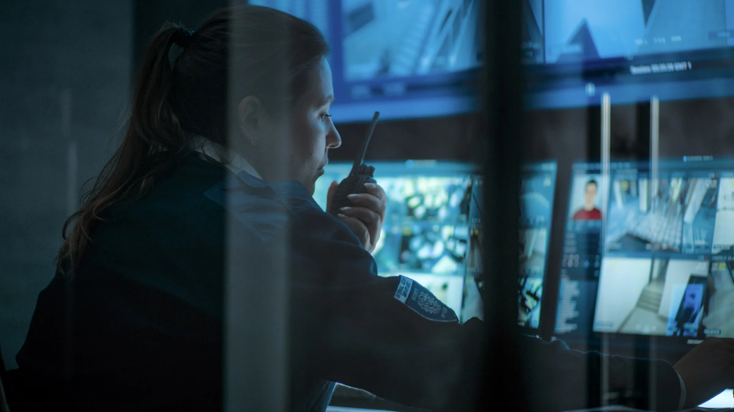 Security officer speaking into a handheld radio by a window at night, with a camera feed visible