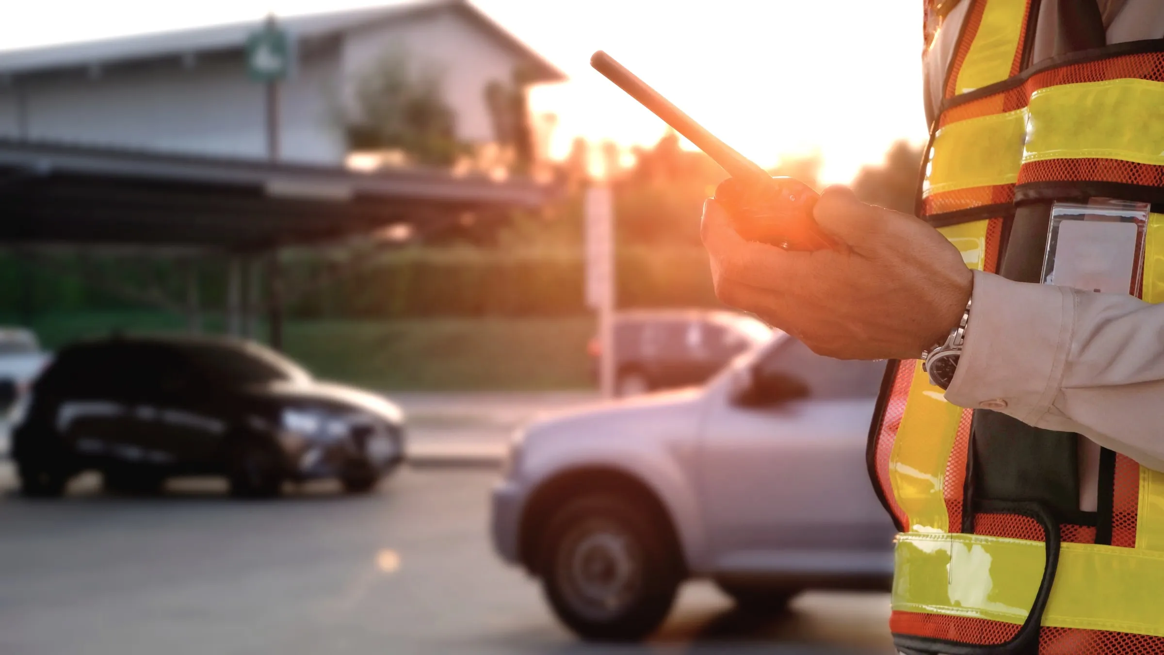 Arm of an officer in a hi-vis vest holding a radio toward a car at a gate in low sun