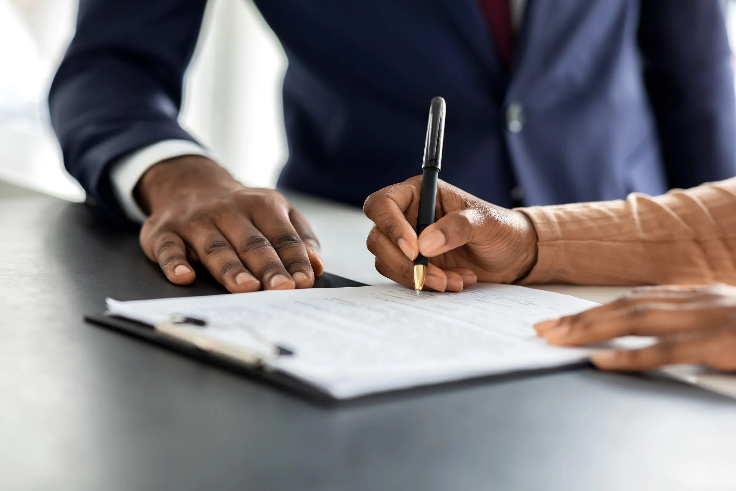 Two people at a desk, one signing a document on a clipboard while the other watches