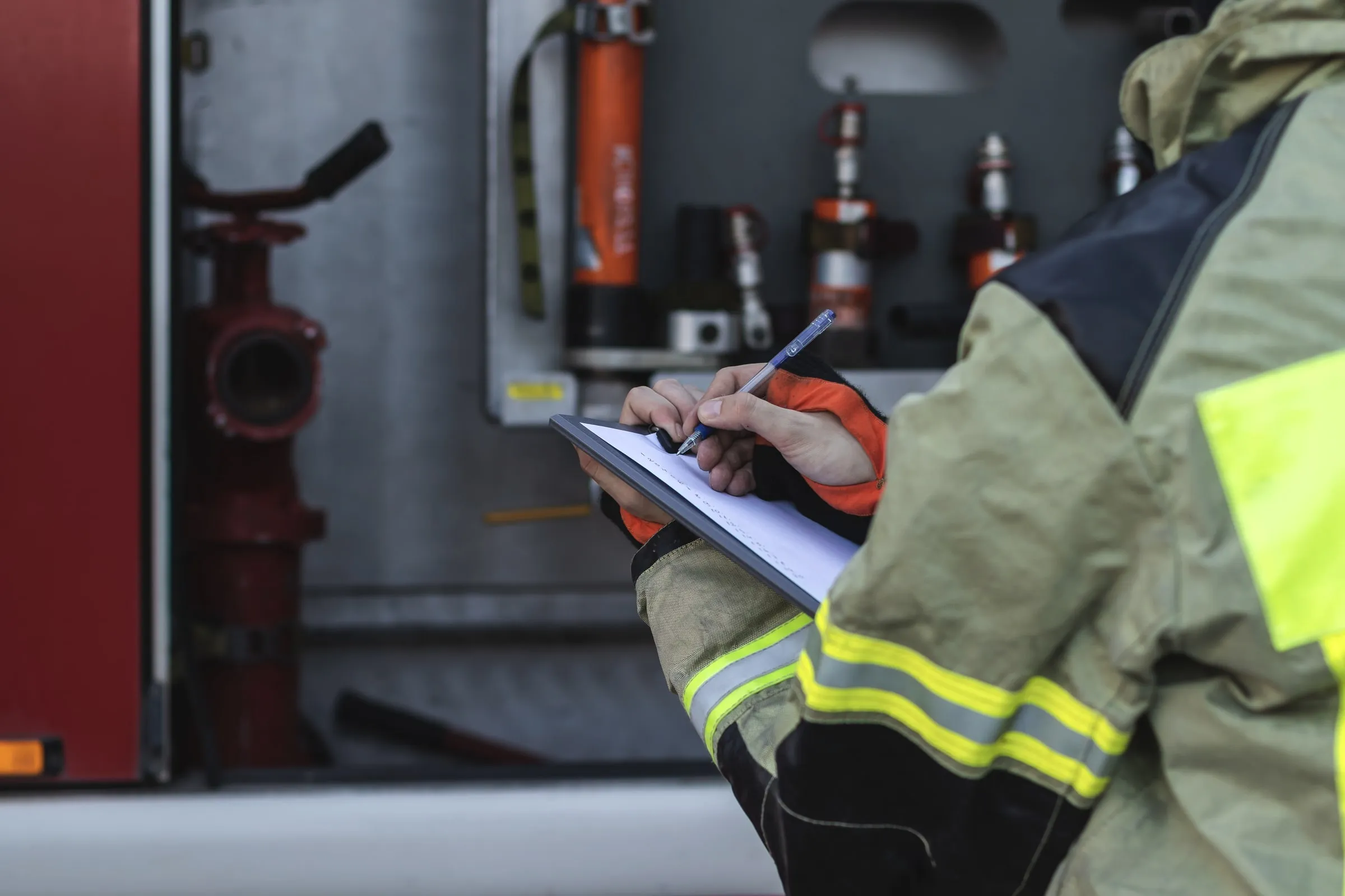 Firefighter in turnout gear writing on a clipboard beside red equipment