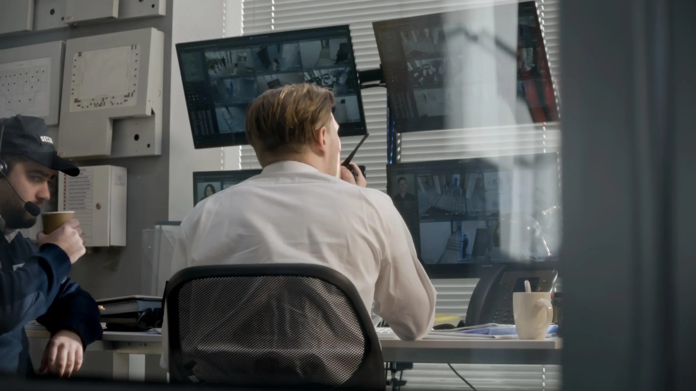 Man in a white shirt seen from behind at a desk, watching a bank of camera monitors