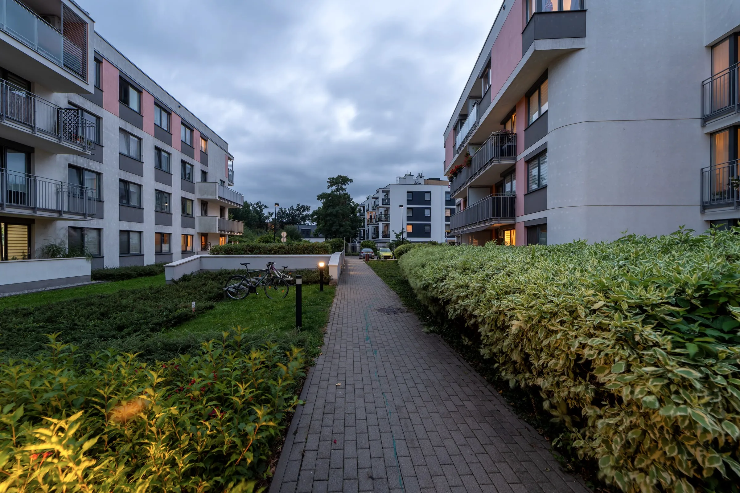 Paved path between landscaped hedges and apartment buildings at dusk under an overcast sky