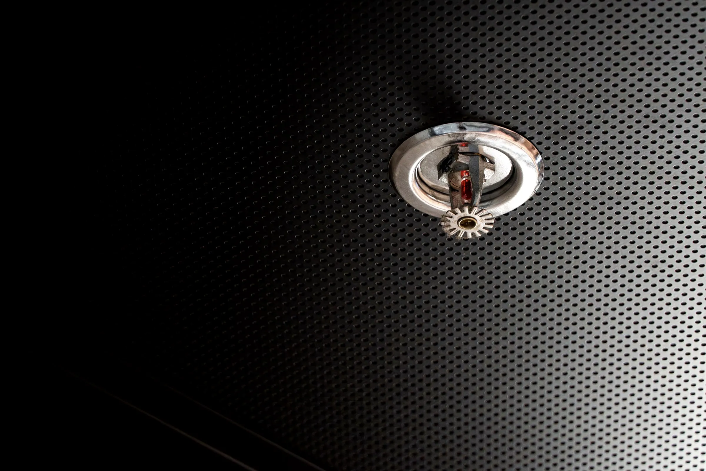 Chrome fire sprinkler head set into a dark perforated ceiling panel