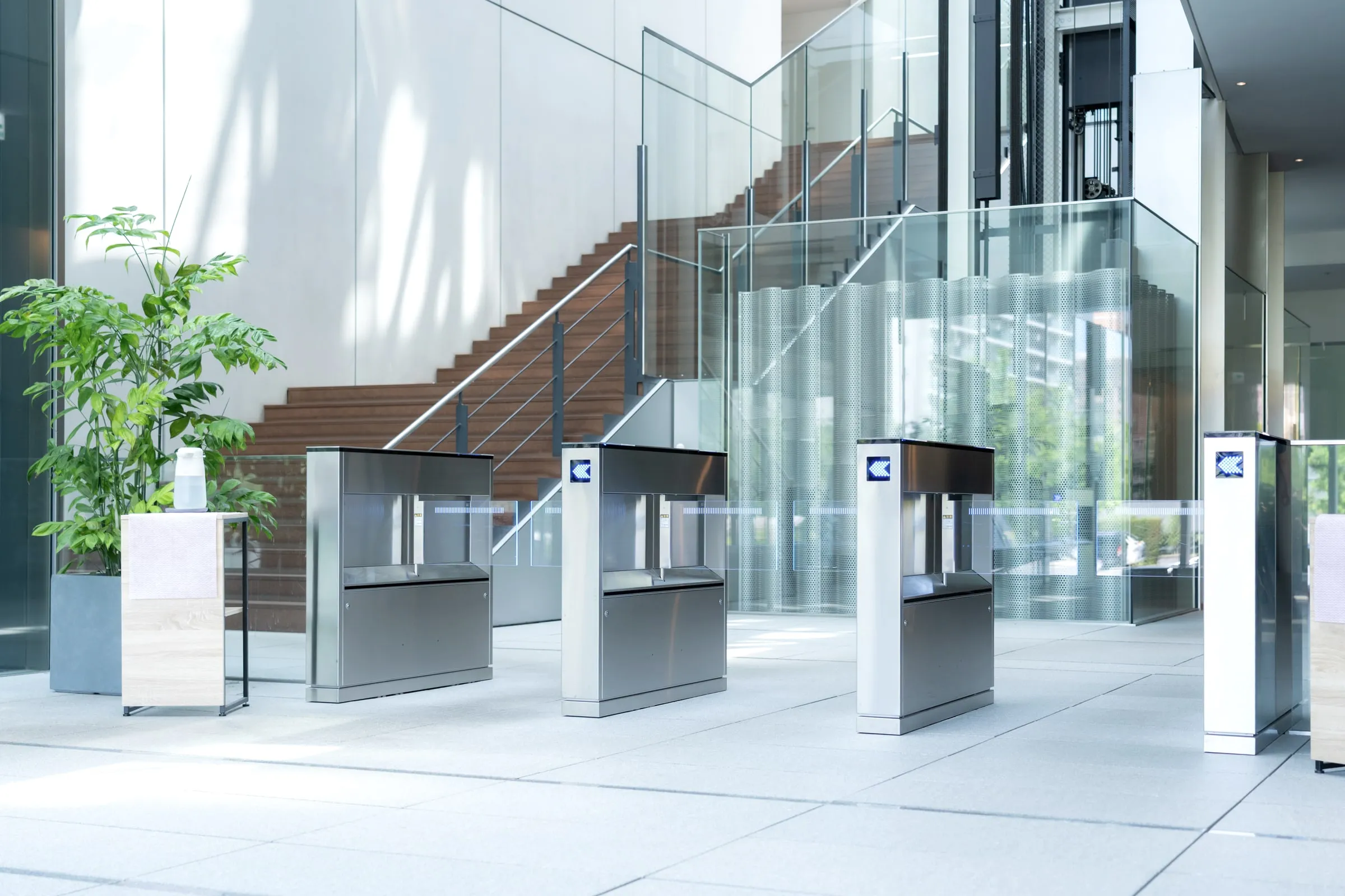 Row of glass security turnstiles in a modern building lobby with plants and stairs behind