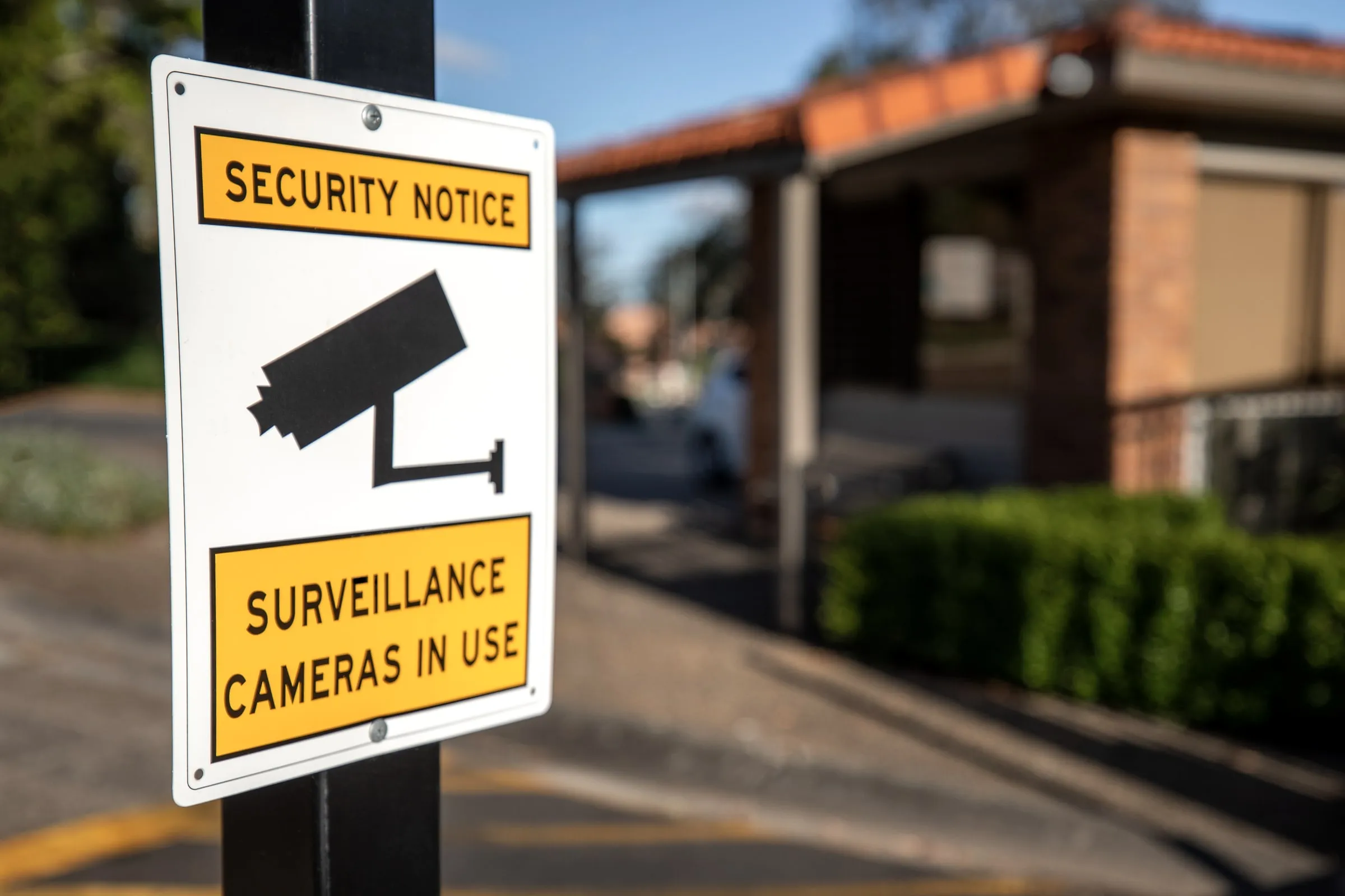 Yellow and black sign reading SECURITY NOTICE, SURVEILLANCE CAMERAS IN USE beside a guardhouse