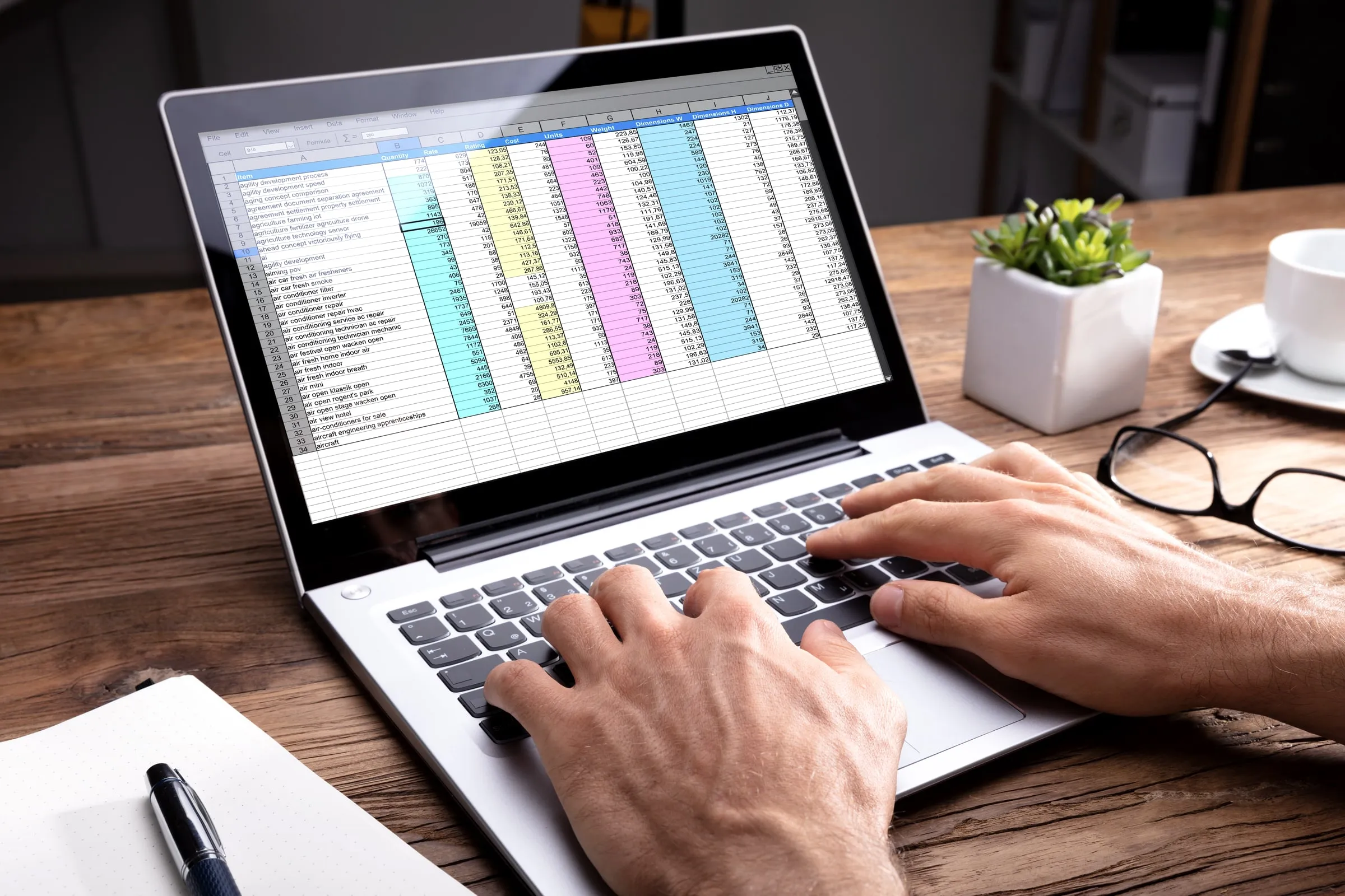 Laptop showing a colorful spreadsheet on a desk beside glasses, a notebook and a small plant