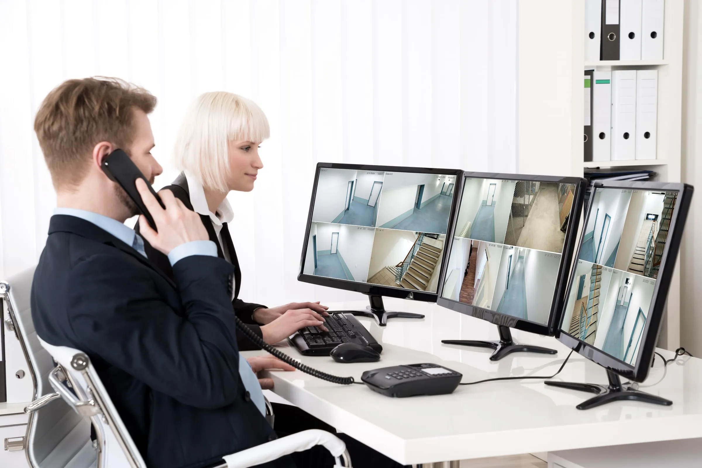 Two security operators at a desk with camera monitors, one speaking on a phone