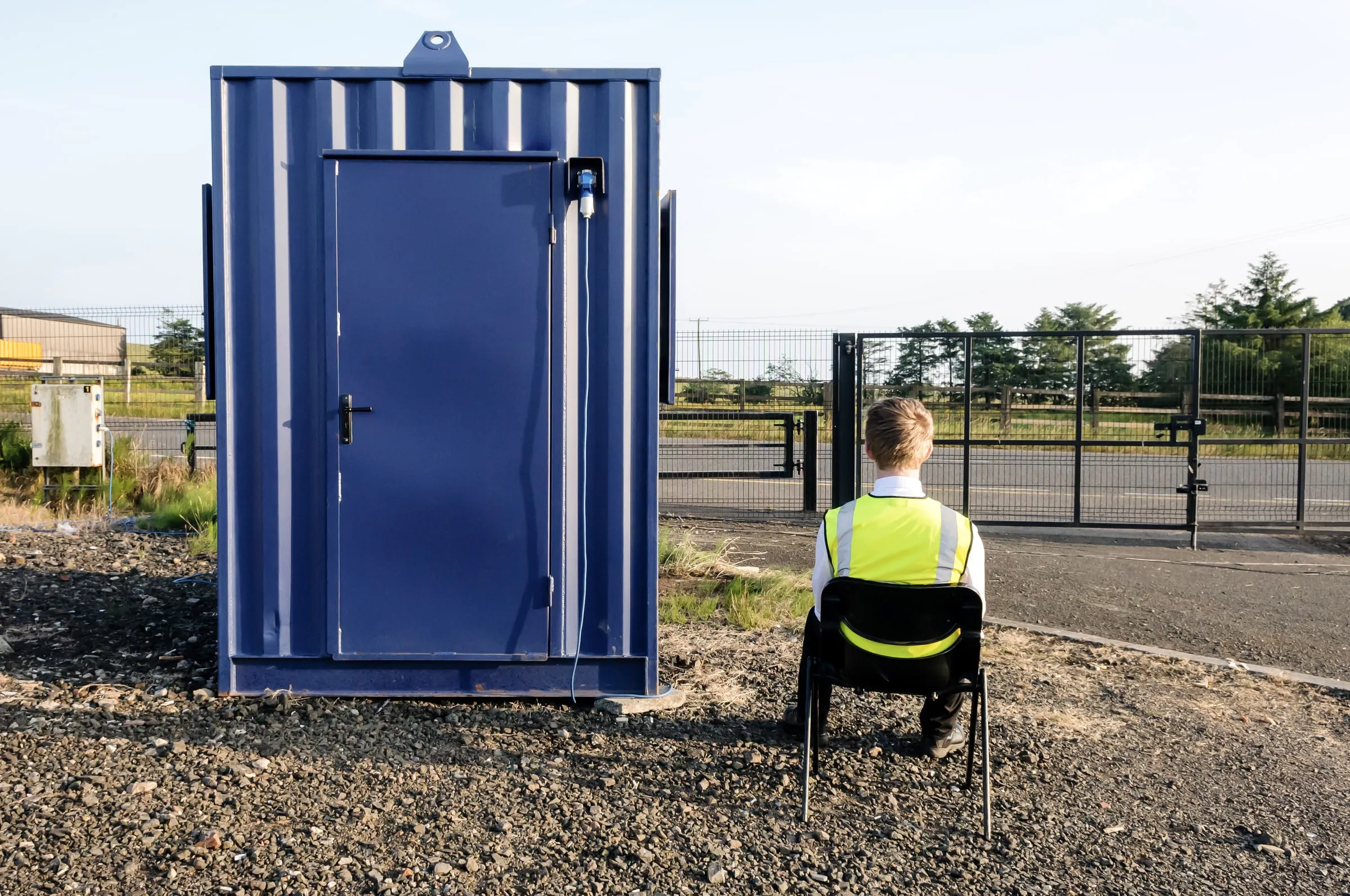 Security officer in a hi-vis vest seated on a chair beside a blue shipping container at a fenced yard gate