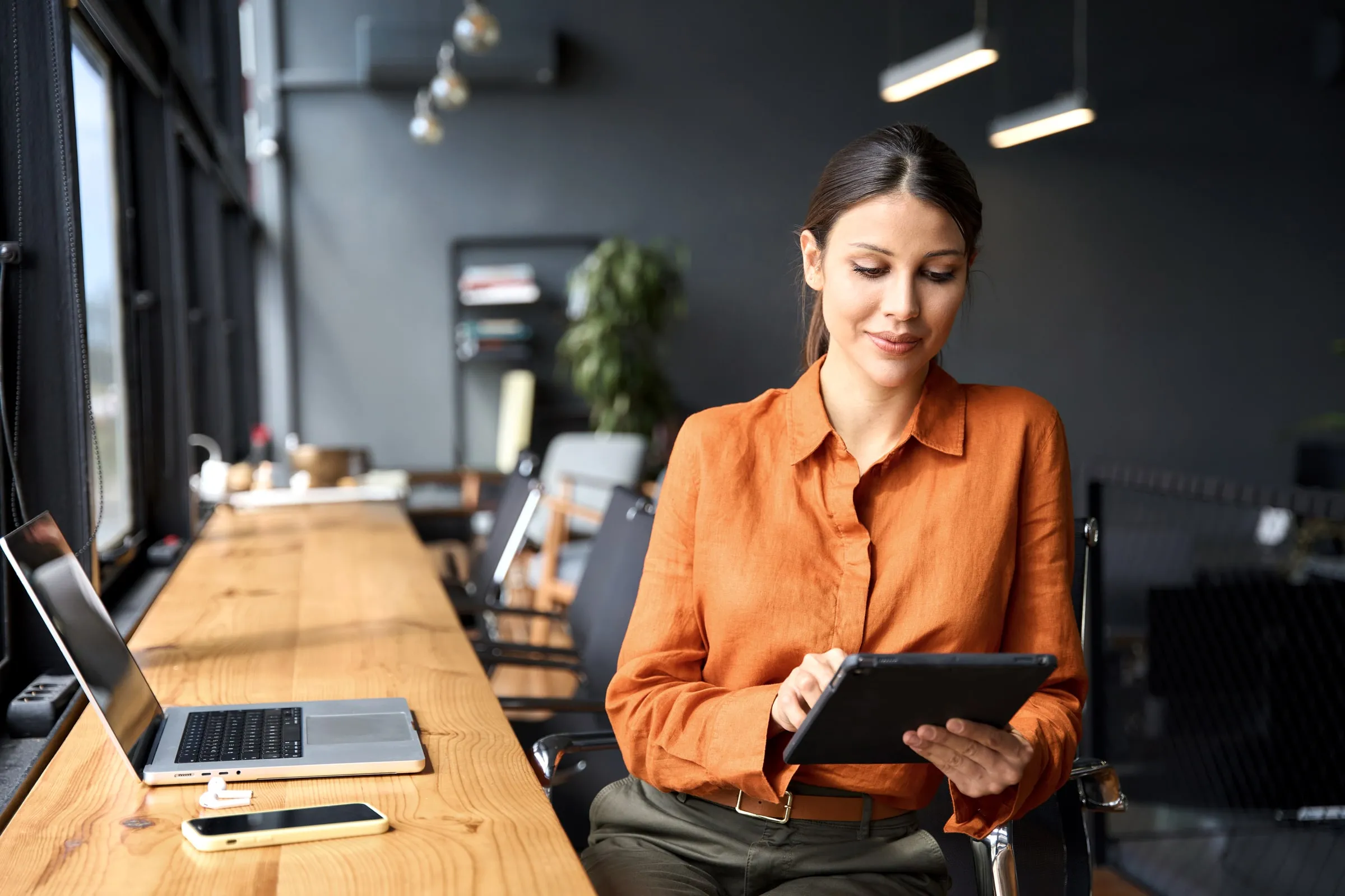 Woman in an orange blouse reading a tablet at a desk in a modern office