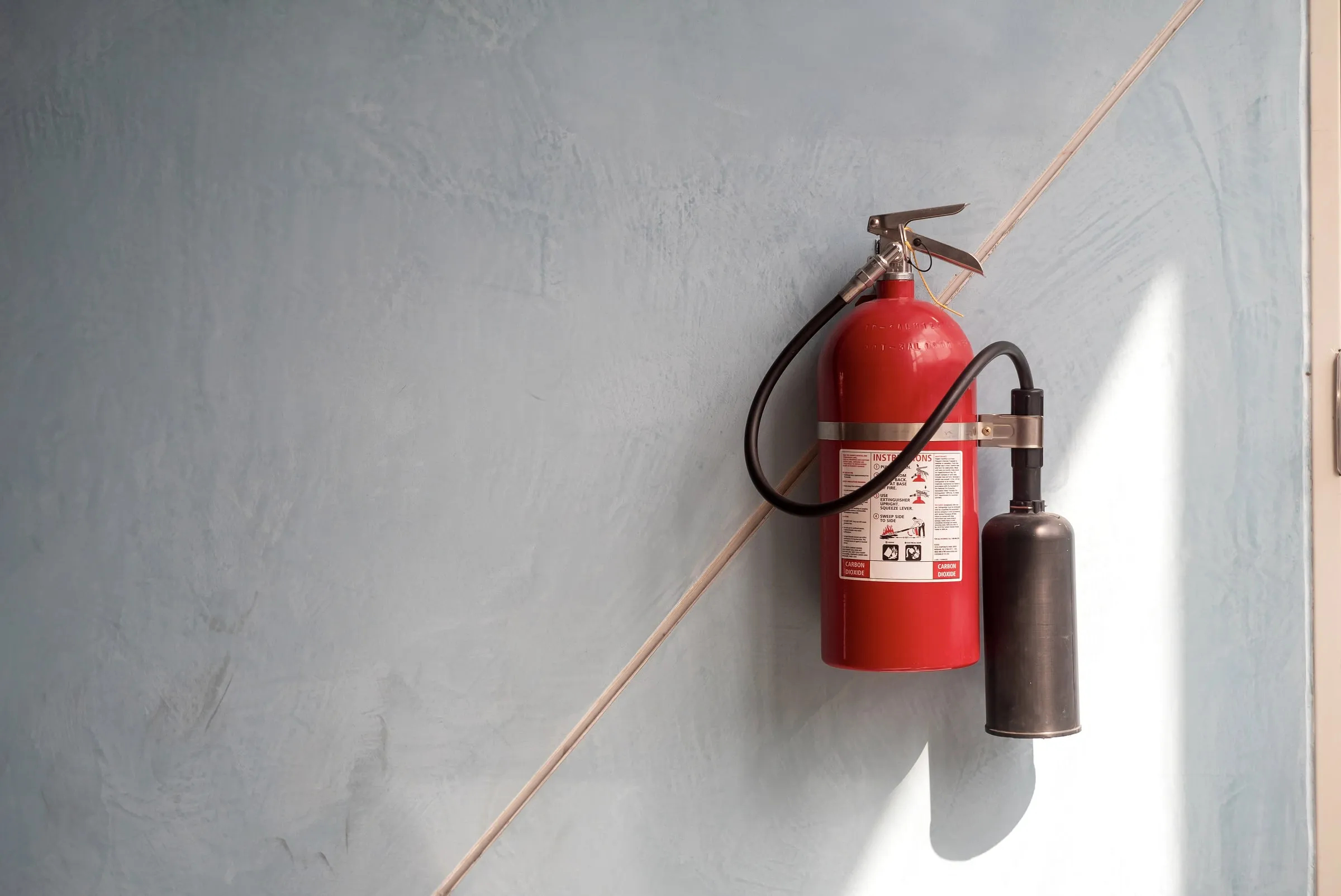 Red fire extinguisher mounted on a grey wall beside a thin pipe