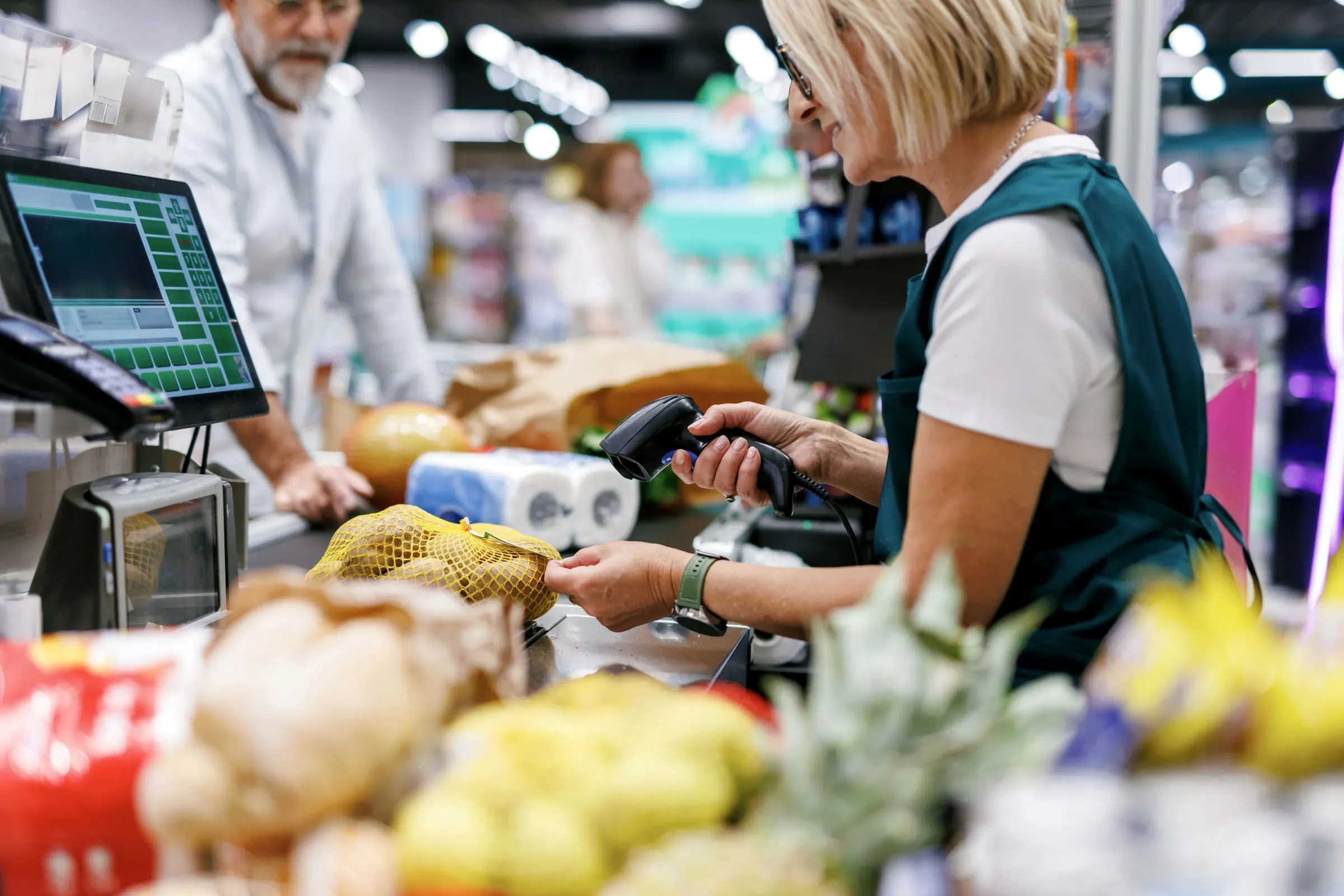 Cashier in a green apron scanning groceries at a busy checkout with customers waiting