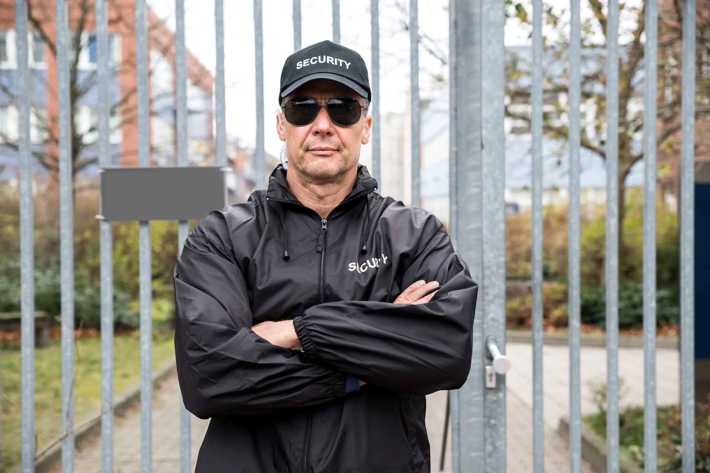 Security officer in a black jacket and cap standing with arms crossed in front of a metal fence