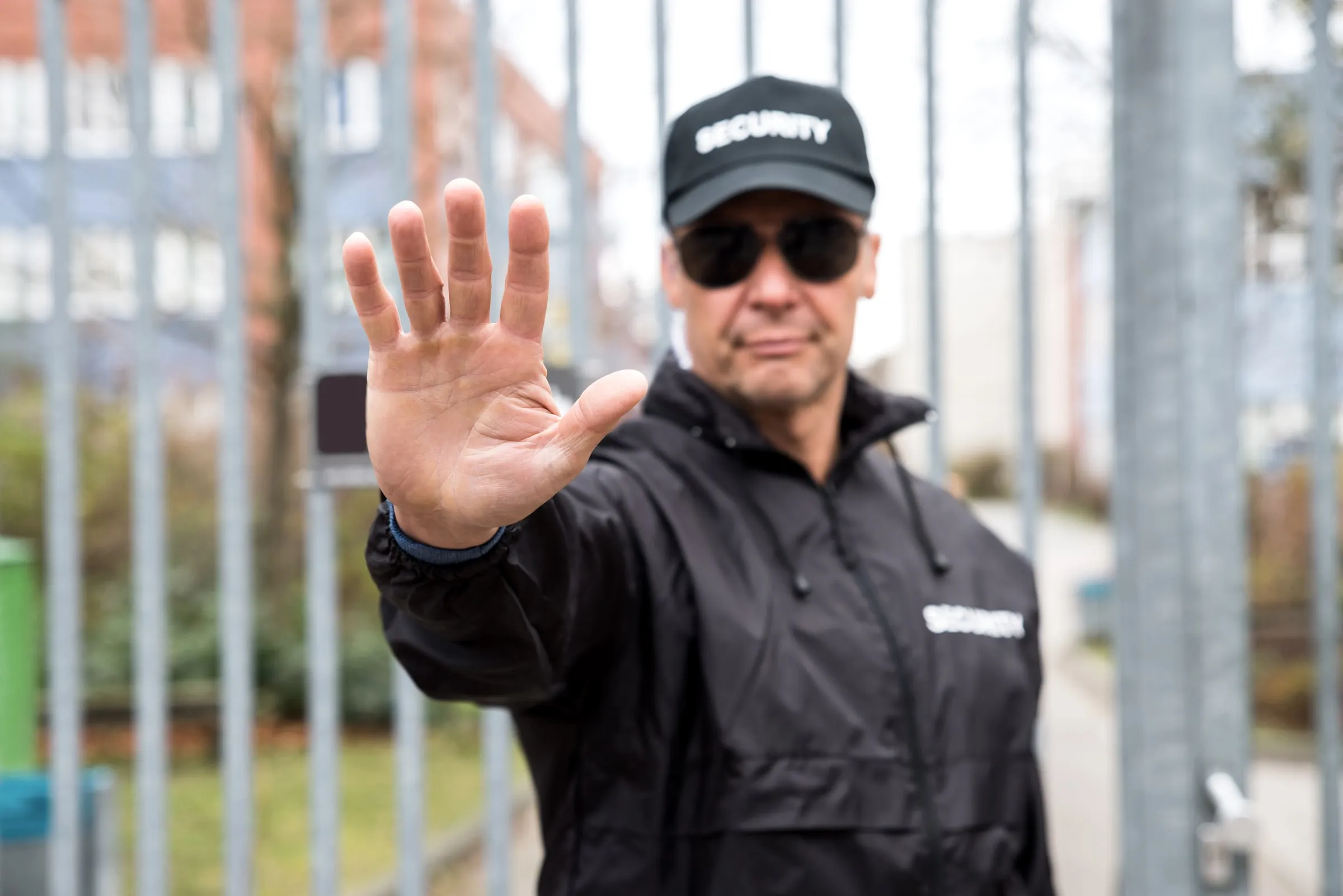 Security officer in a black jacket and cap raising an open palm toward the camera in a fenced yard