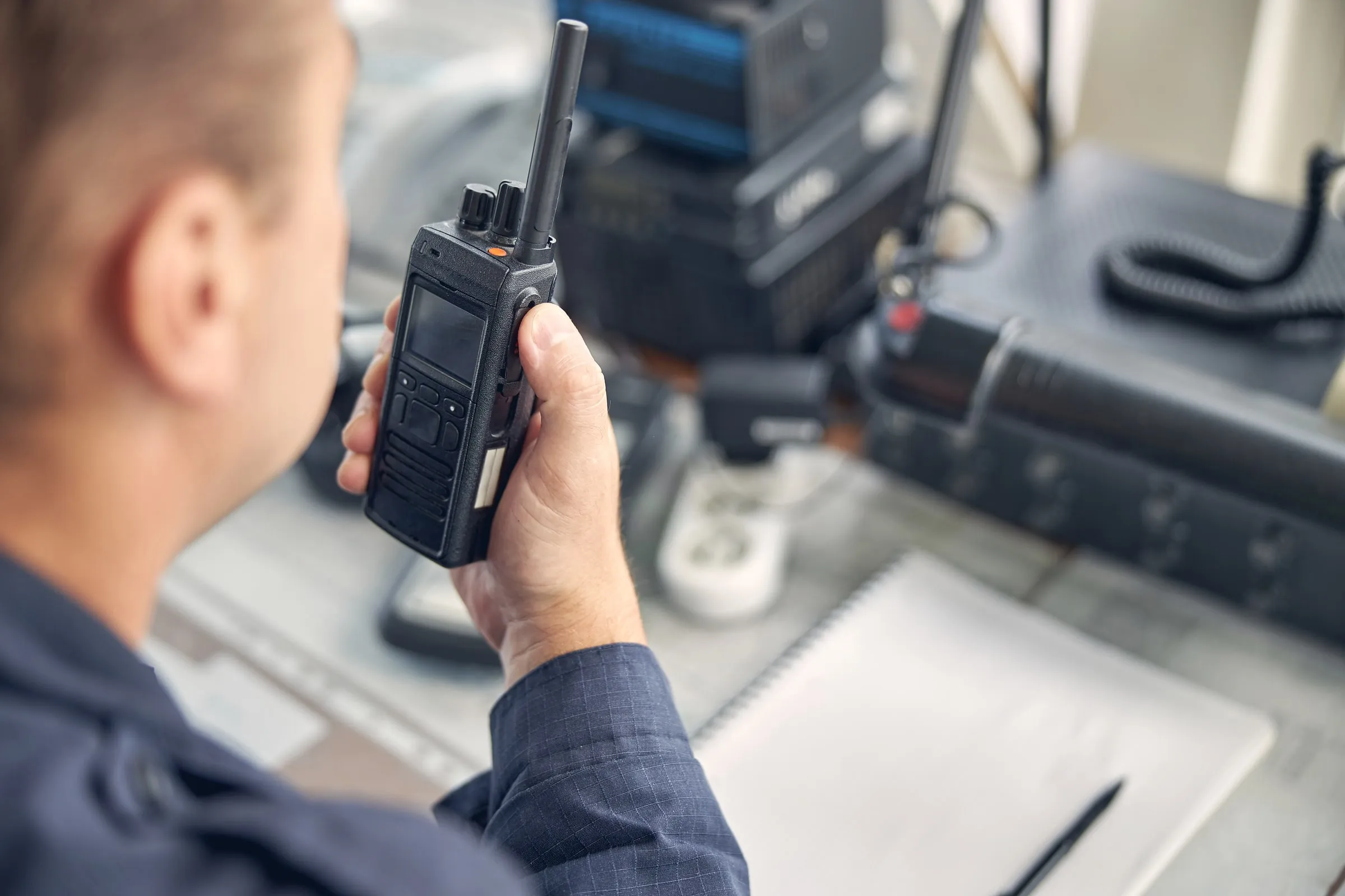 Security officer in a dark uniform holding a handheld radio in front of a control desk