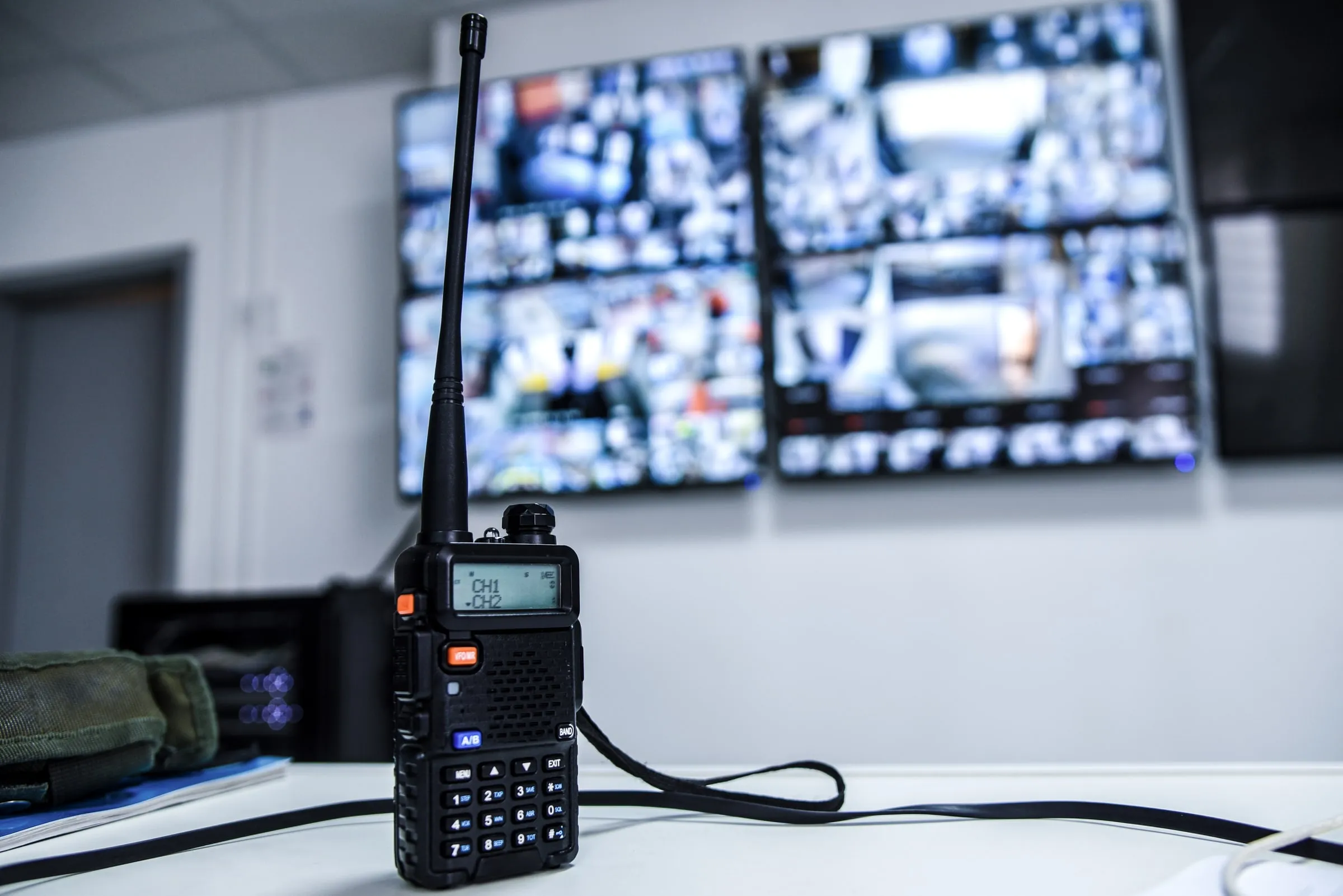 Handheld radio standing on a desk in front of a wall of camera monitors