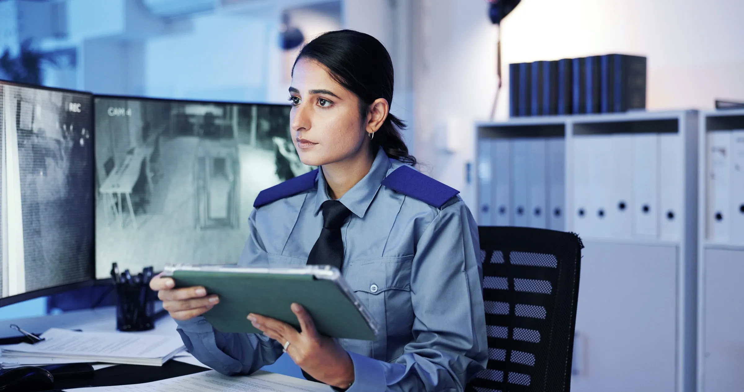 Female security officer in a blue uniform reading a tablet at a desk in a control room