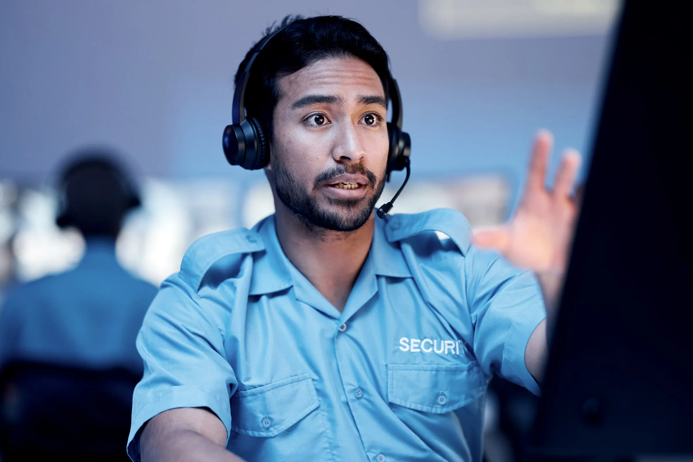 Security officer in a blue uniform and headset gesturing at a screen in a control room