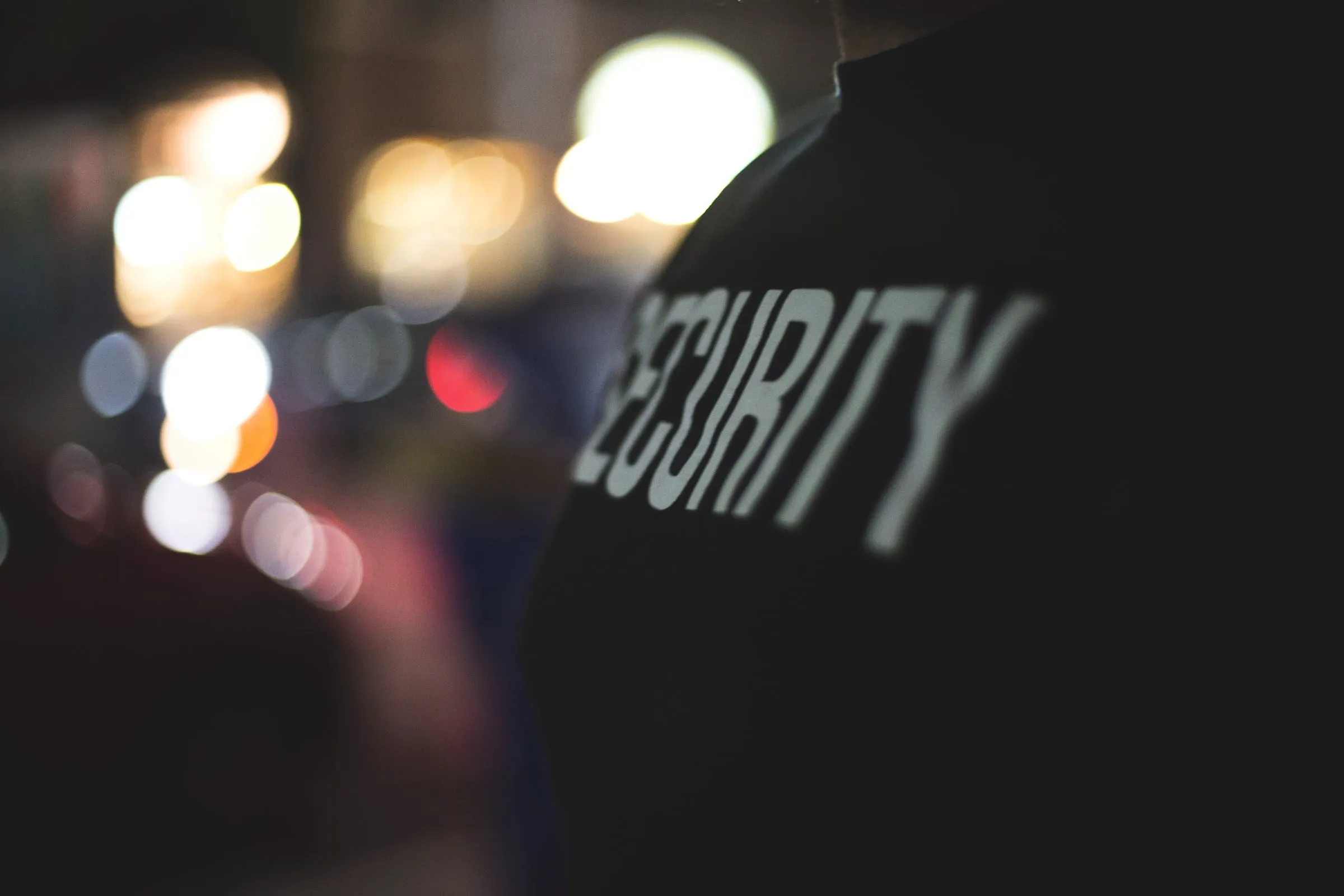 Back of a security officer's dark jacket with SECURITY lettering against blurred city lights at night