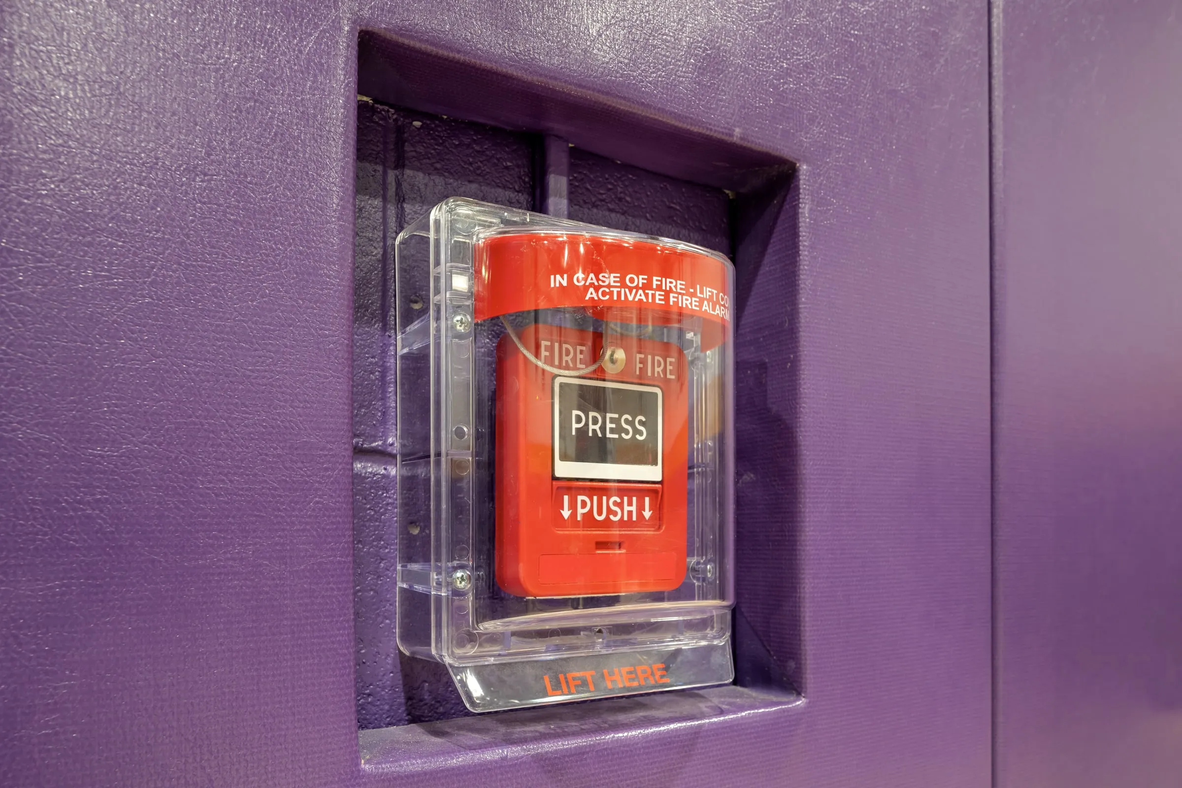 Red fire alarm call point with a clear cover on a purple wall