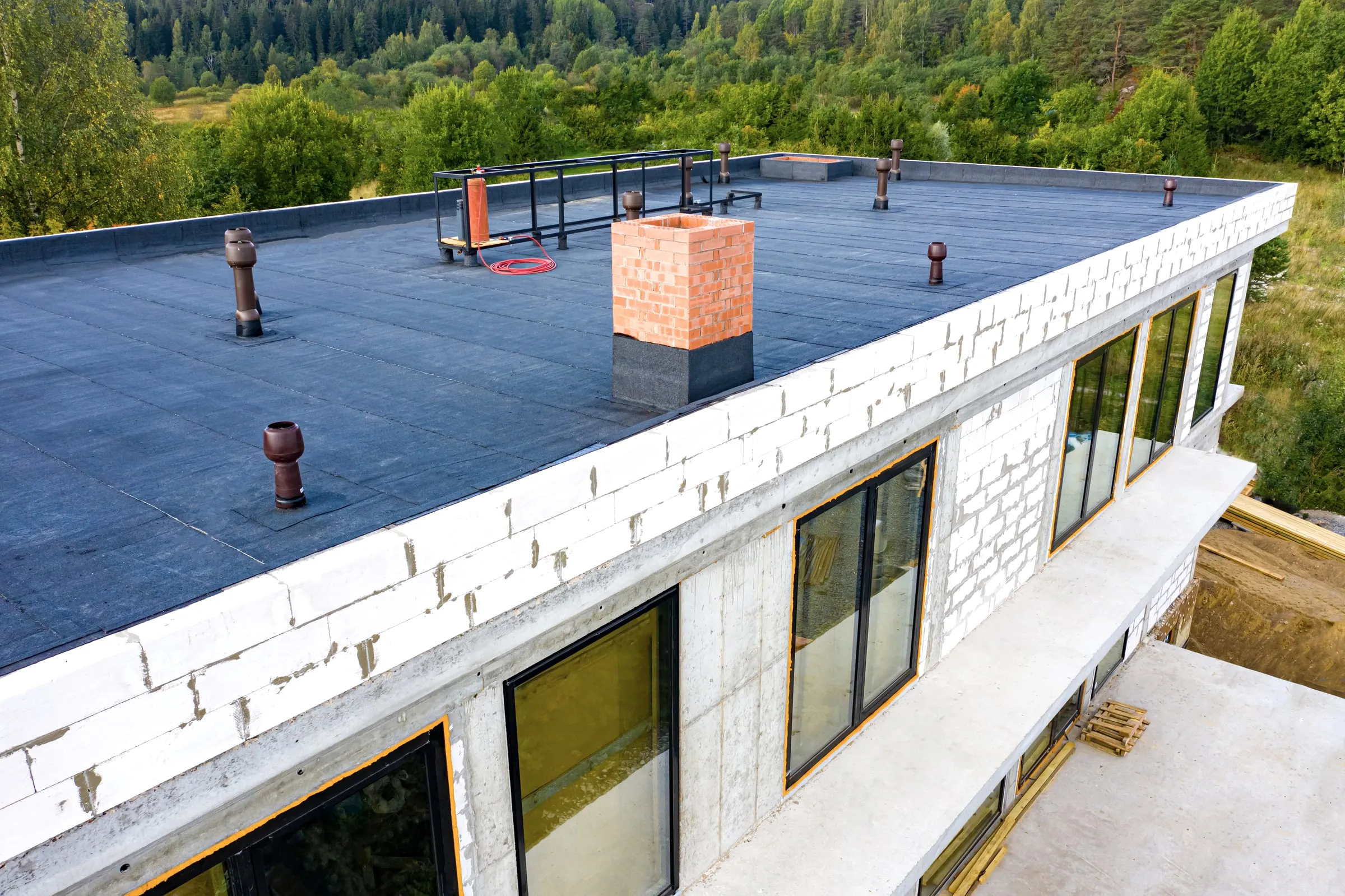 Flat black roof with a parapet, vents and a chimney, seen from above with trees beyond