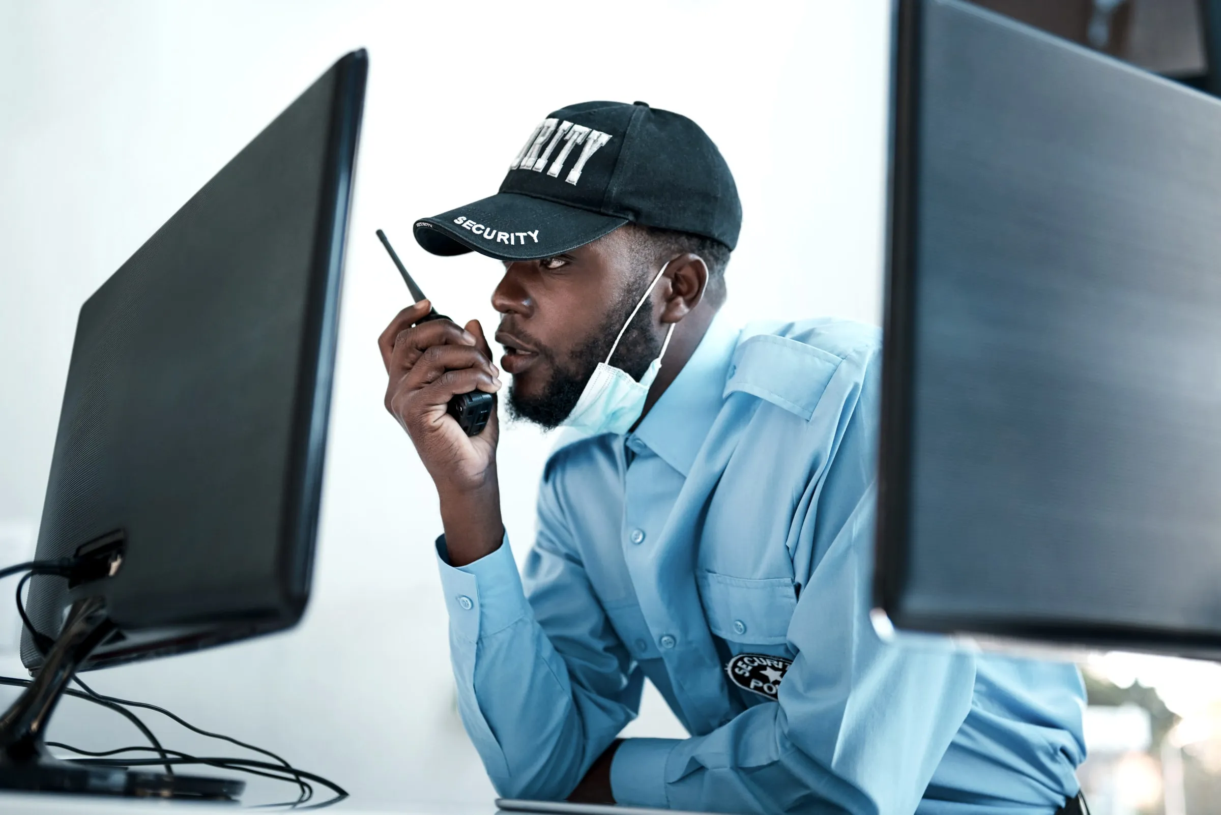 Commissioned security officer in a light blue uniform and SECURITY cap speaking into a radio between two monitors