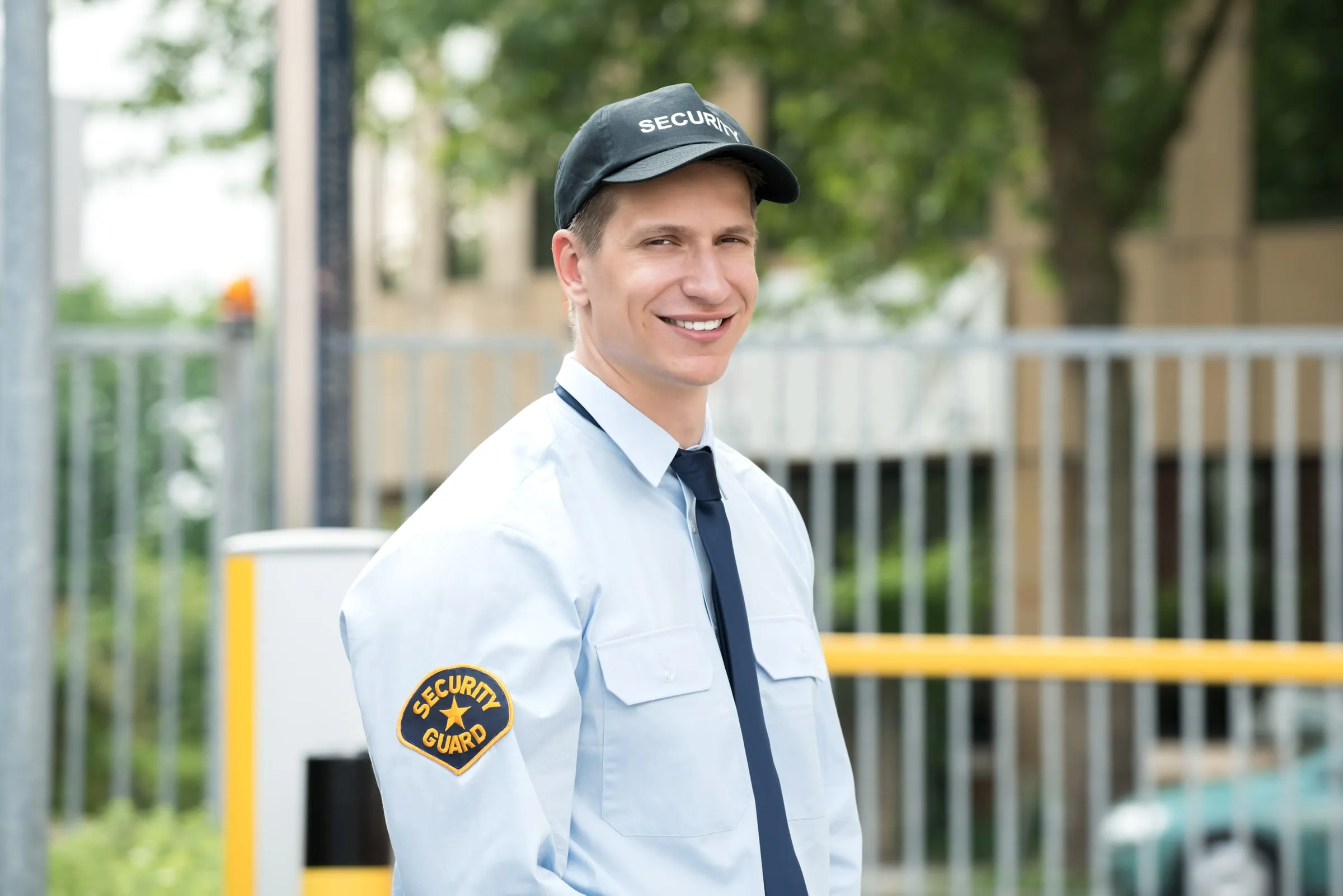 Smiling security officer in a white uniform shirt and SECURITY cap standing beside a gate arm