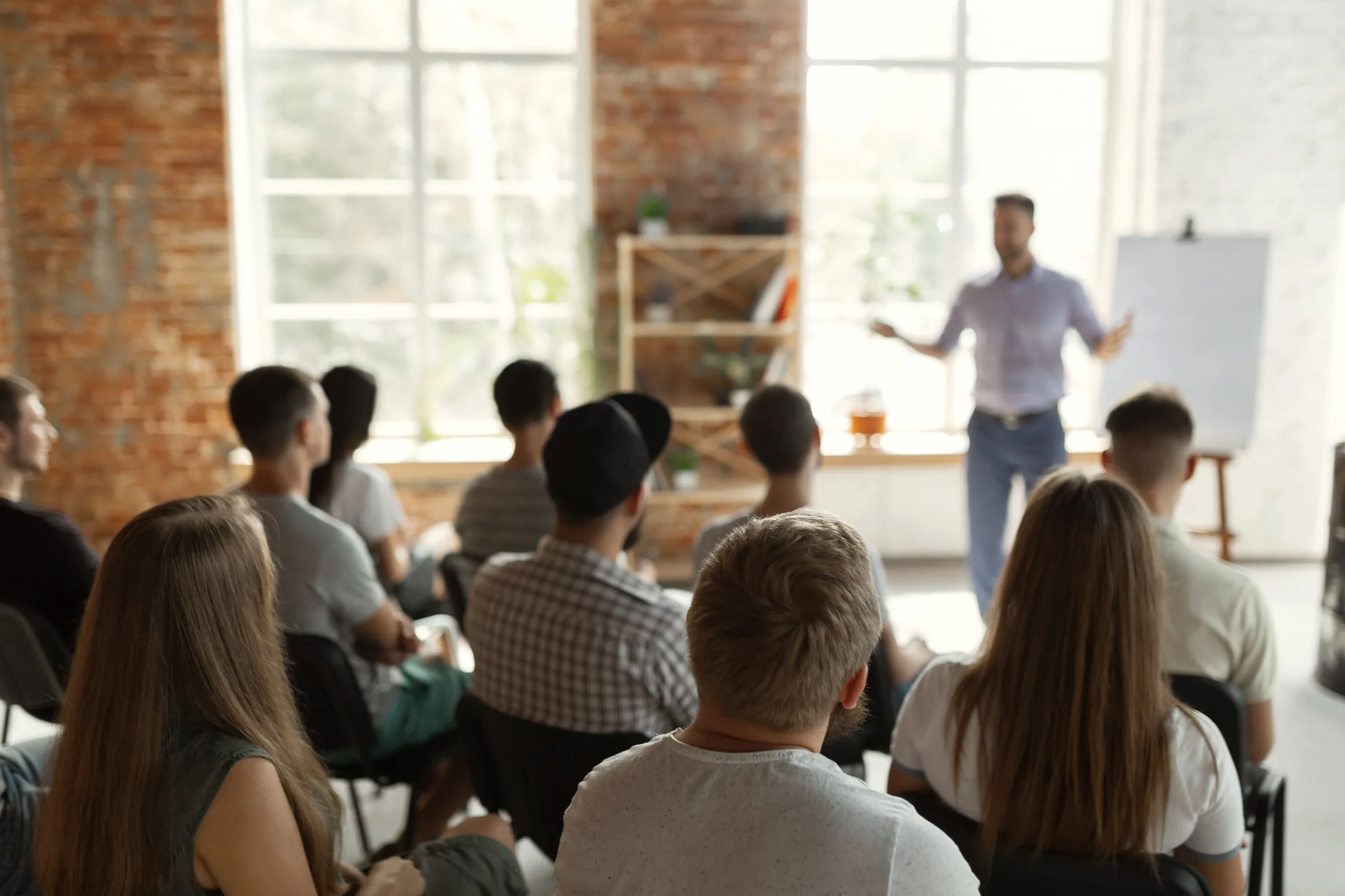Instructor at a whiteboard addressing a seated class in a bright brick-walled training room